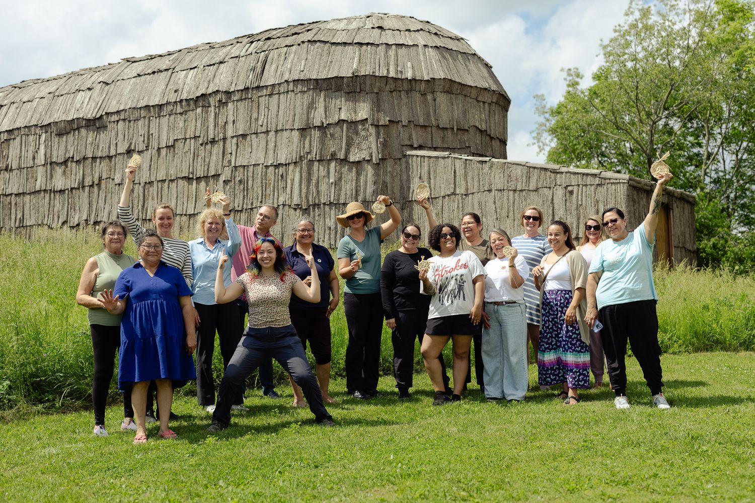 Group of people posing and celebrating outdoors near a traditional Indigenous longhouse structure on a sunny day.