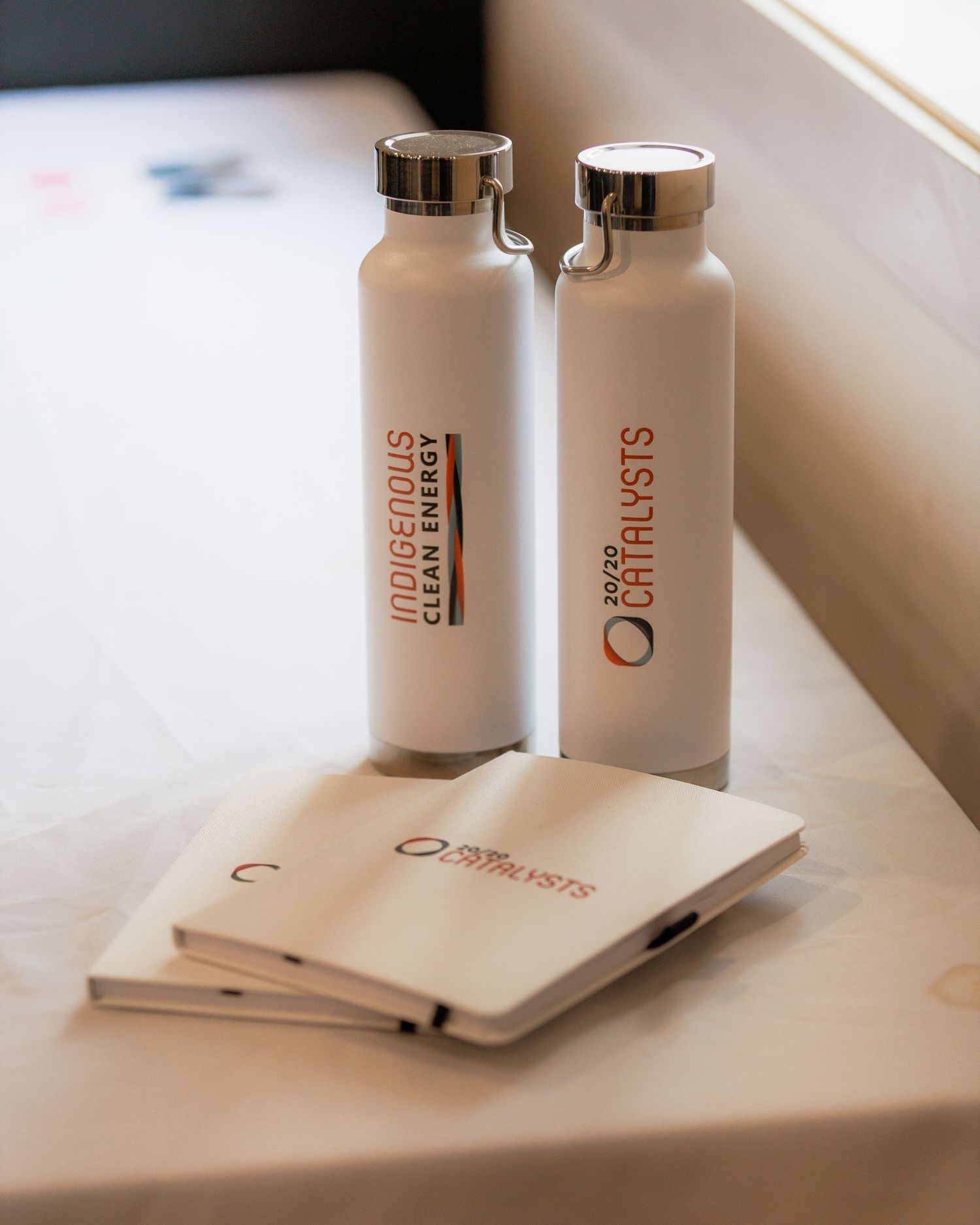 White Indigenous Clean Energy and 2023 Catalysts branded water bottles and notebooks displayed on a white surface.