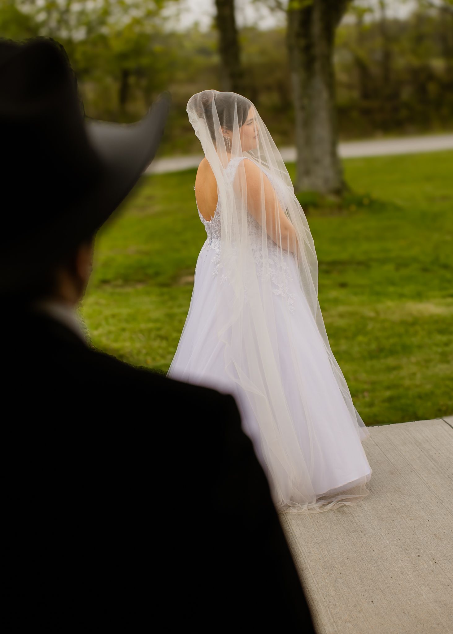 Groom in cowboy hat watches bride in flowing white veil walk away in outdoor setting.