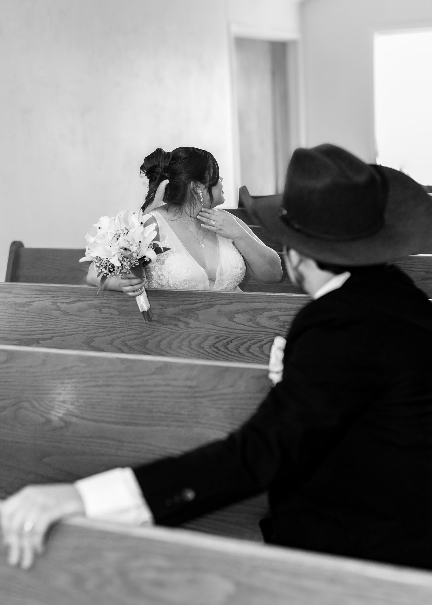 Black and white photo of a bride holding a bouquet in church pews as a groom in a cowboy hat watches.