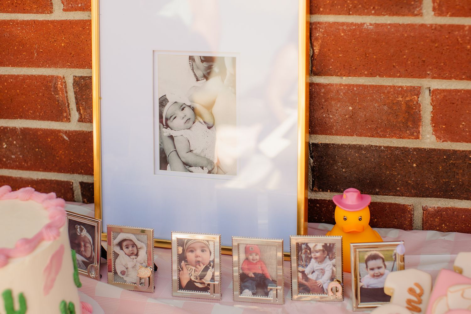 A display of baby photos in gold frames arranged on a table with a cake beside a brick wall.