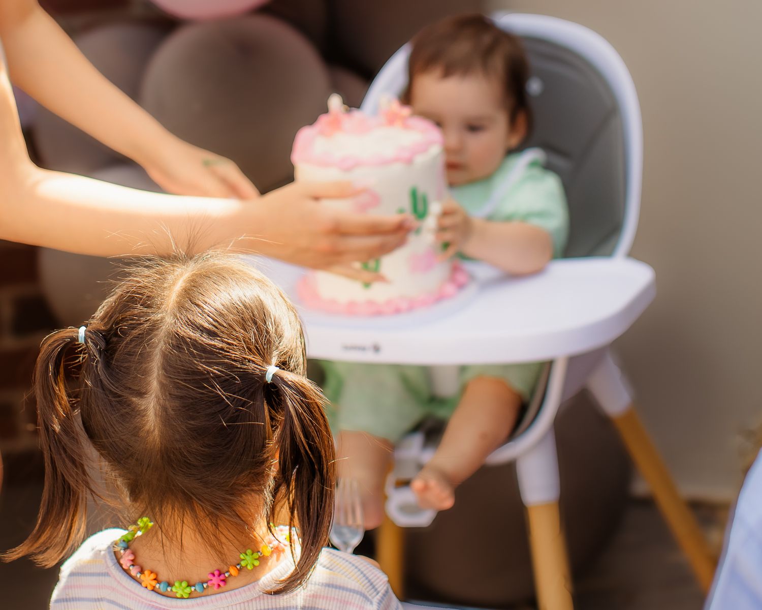 Baby in high chair receives pink first birthday smash cake while toddler watches nearby.