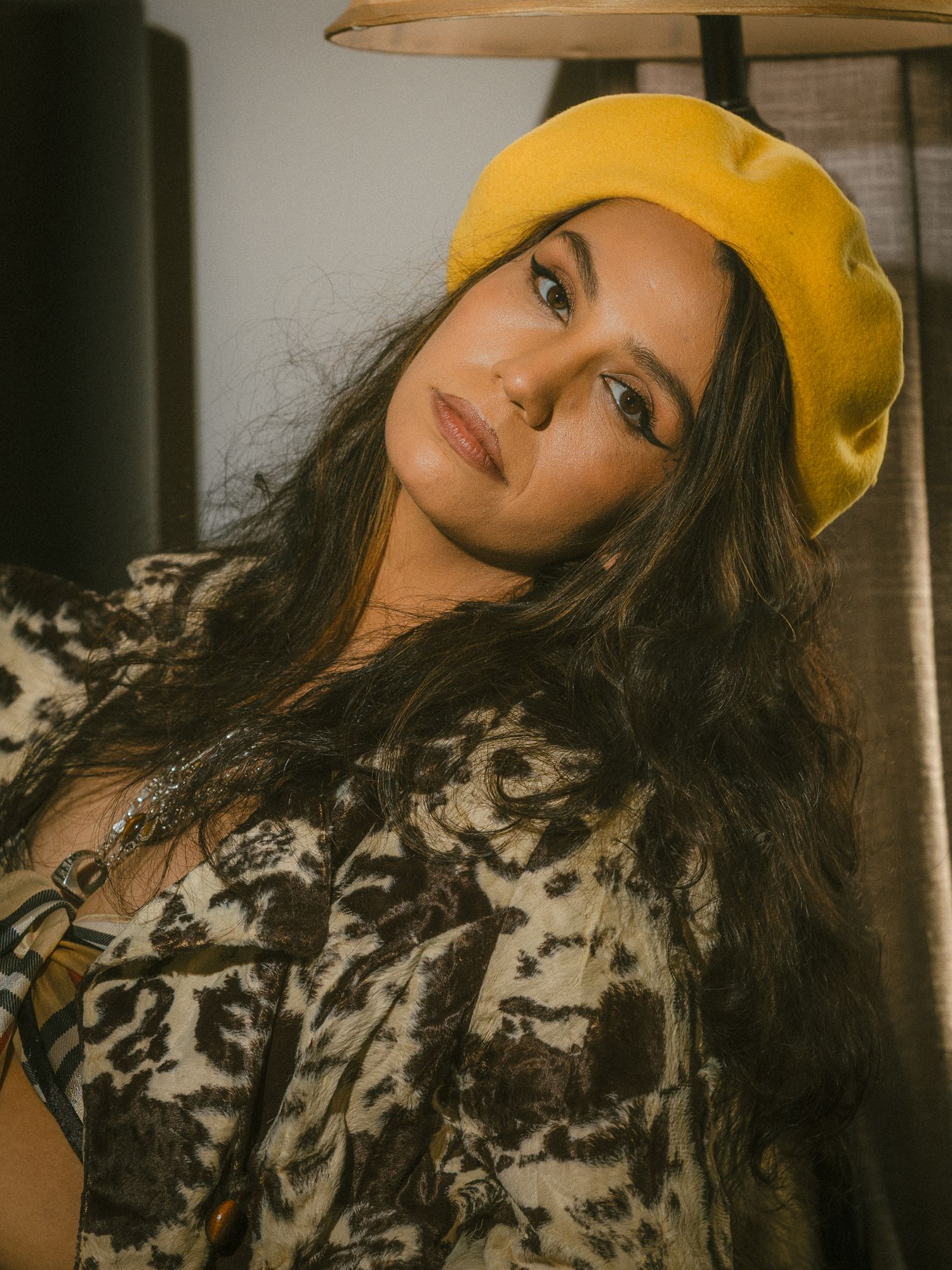 Young woman in yellow beret and animal print jacket posing indoors with long dark wavy hair.
