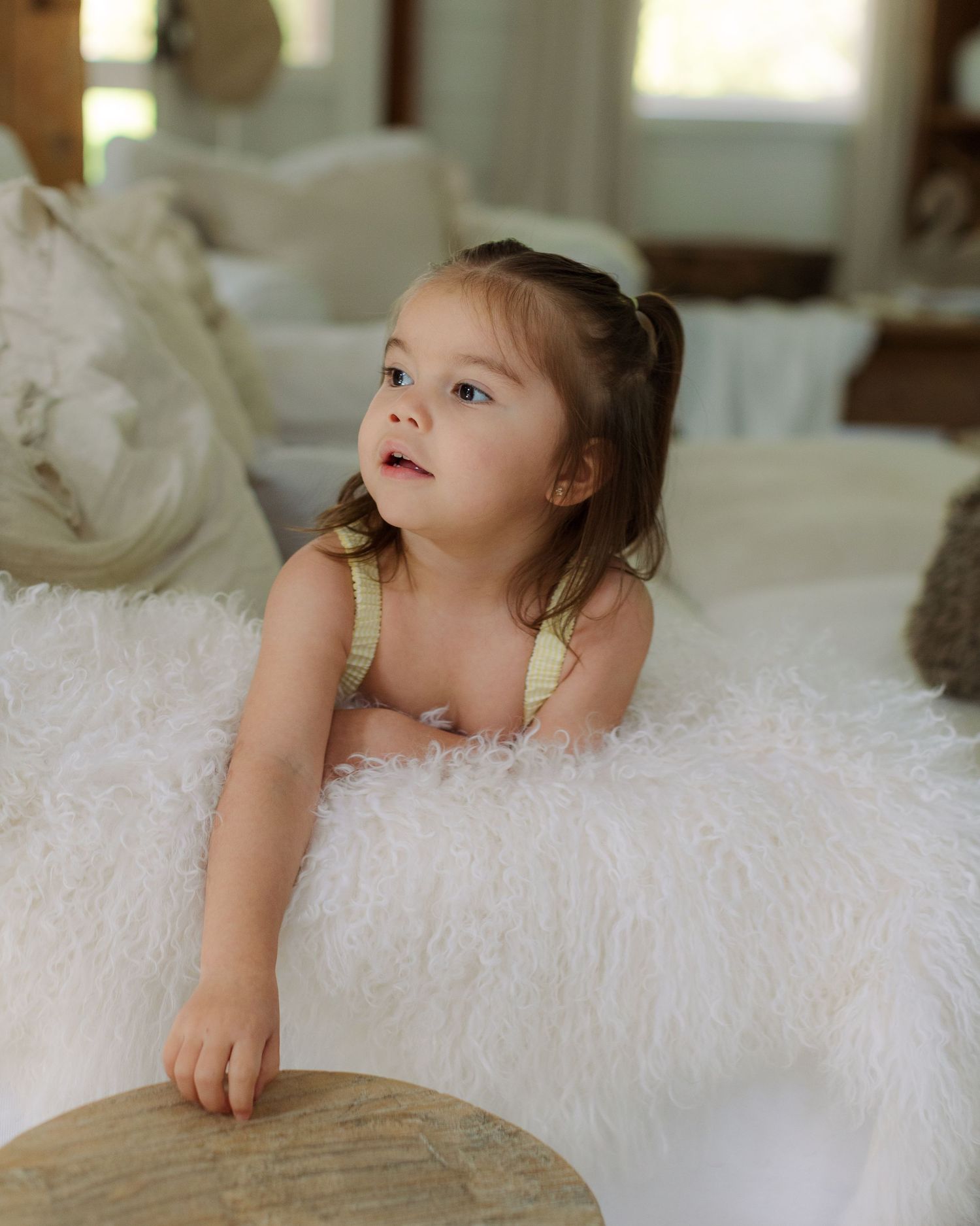 A young toddler girl with pigtails leans over a fluffy white ottoman in a bright, cozy living room.