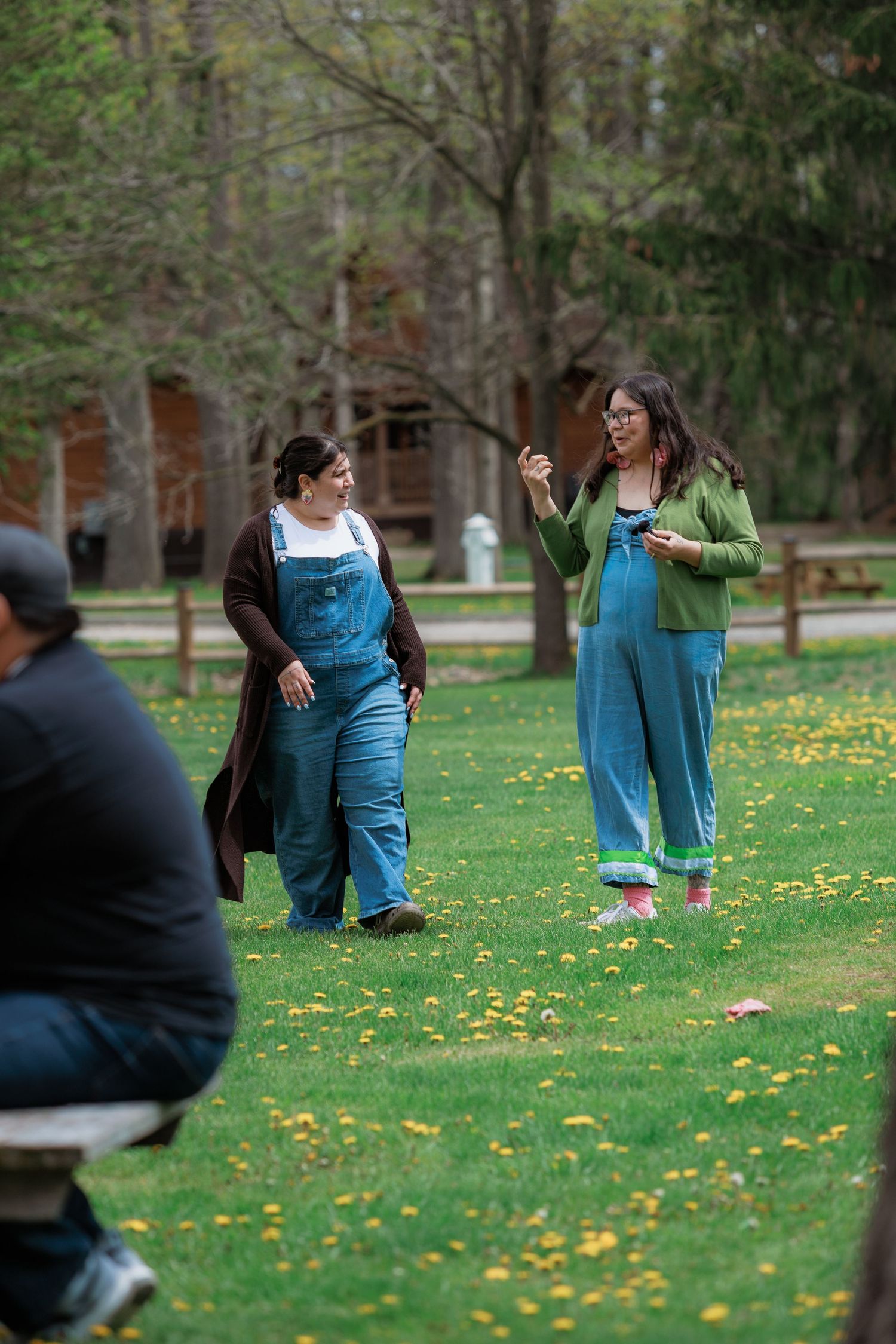 Two women in overalls conversing on green lawn with dandelions in a park setting.