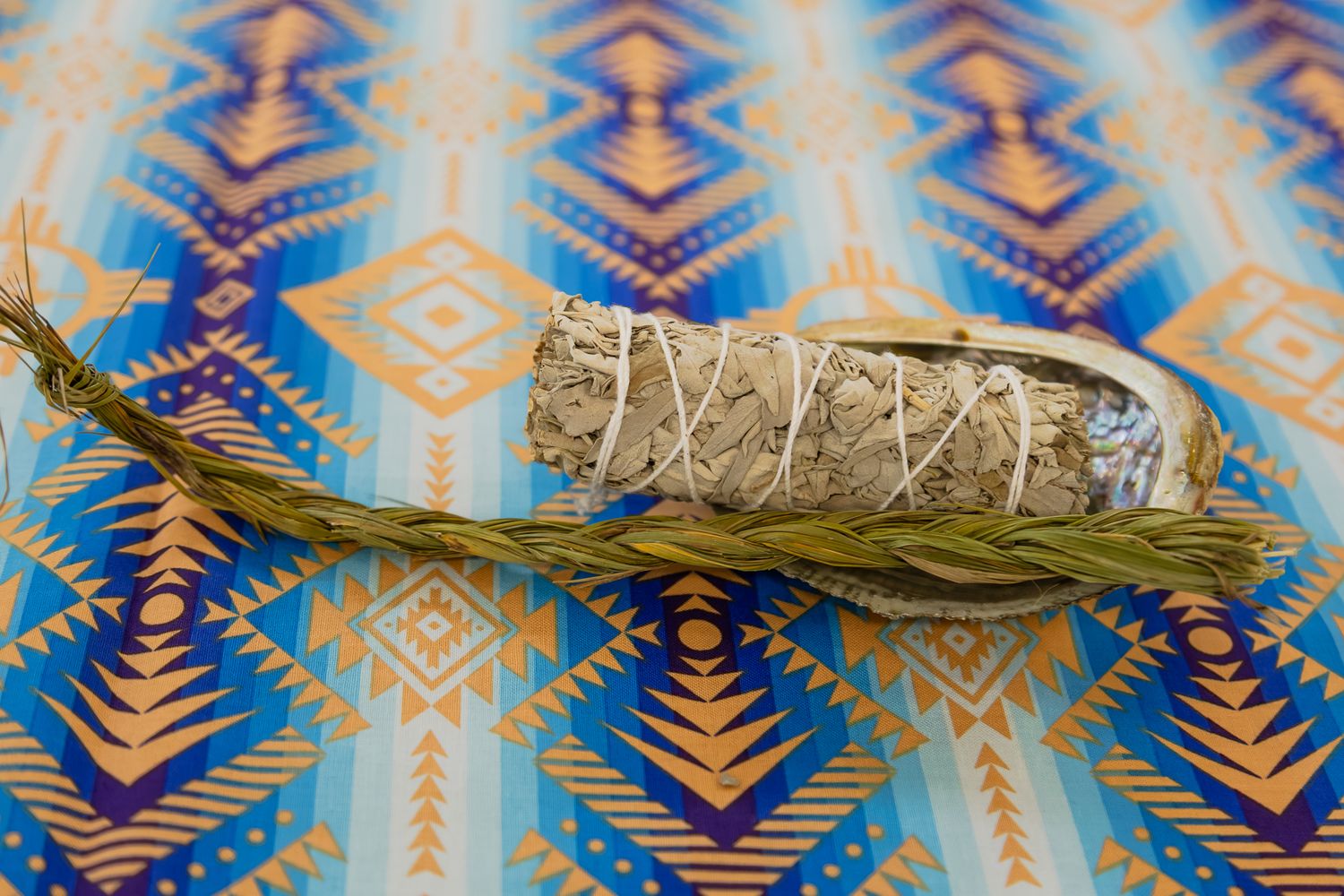 A white sage smudge stick with dried grass on a blue and gold Native American geometric patterned fabric background.
