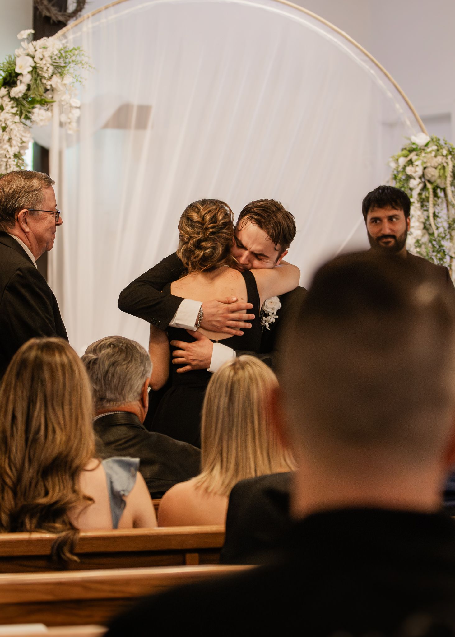 Groom hugging a guest in black dress at wedding ceremony with floral arch backdrop.