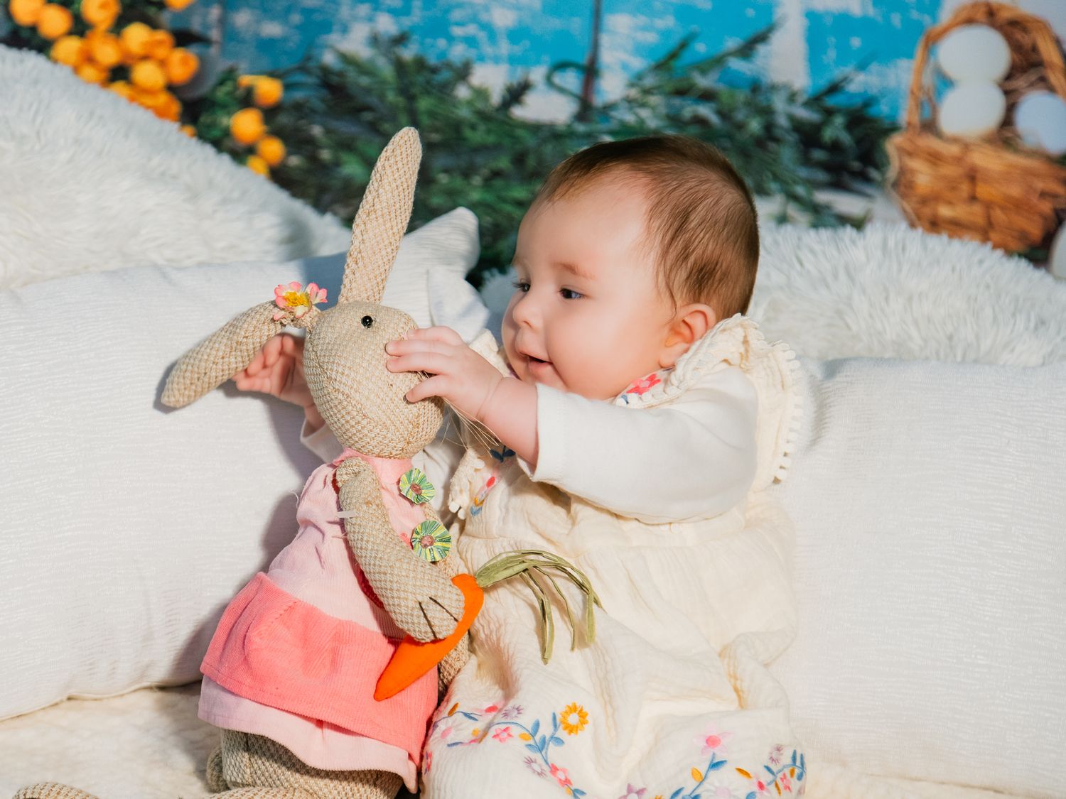 Baby in white dress playing with a stuffed bunny toy on a white couch with Easter decorations in background.