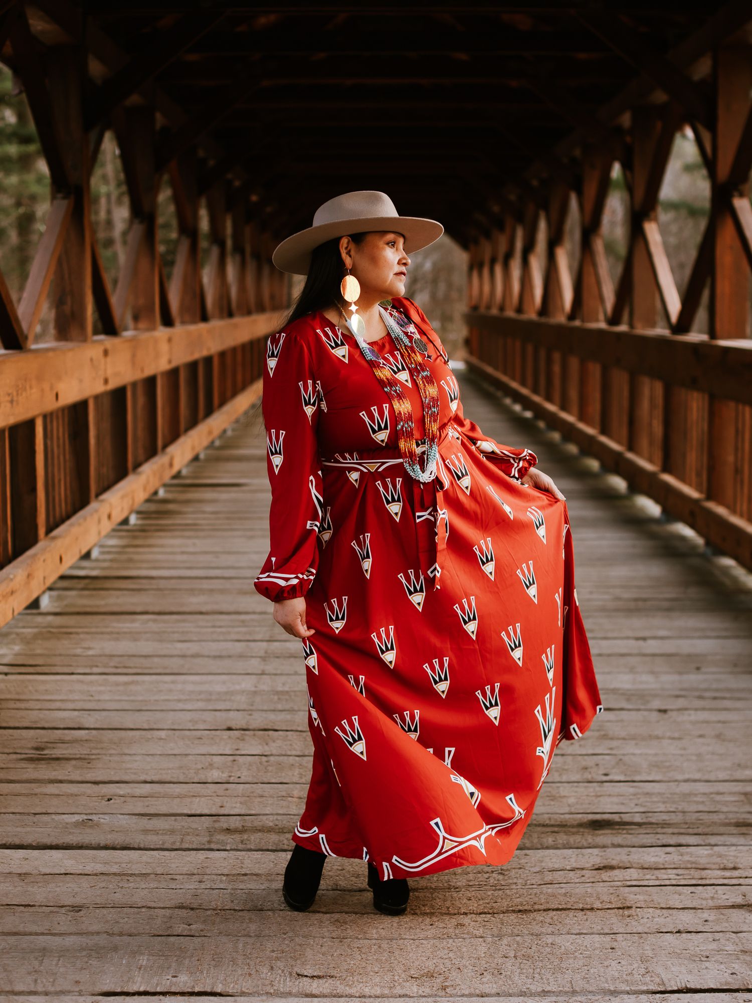 Woman in flowing red patterned dress and tan hat poses on a rustic wooden covered bridge.