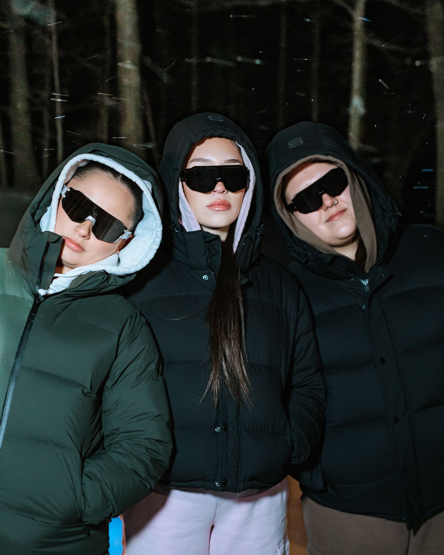 Three women in dark hooded puffer jackets and sunglasses posing outdoors in a snowy forest.