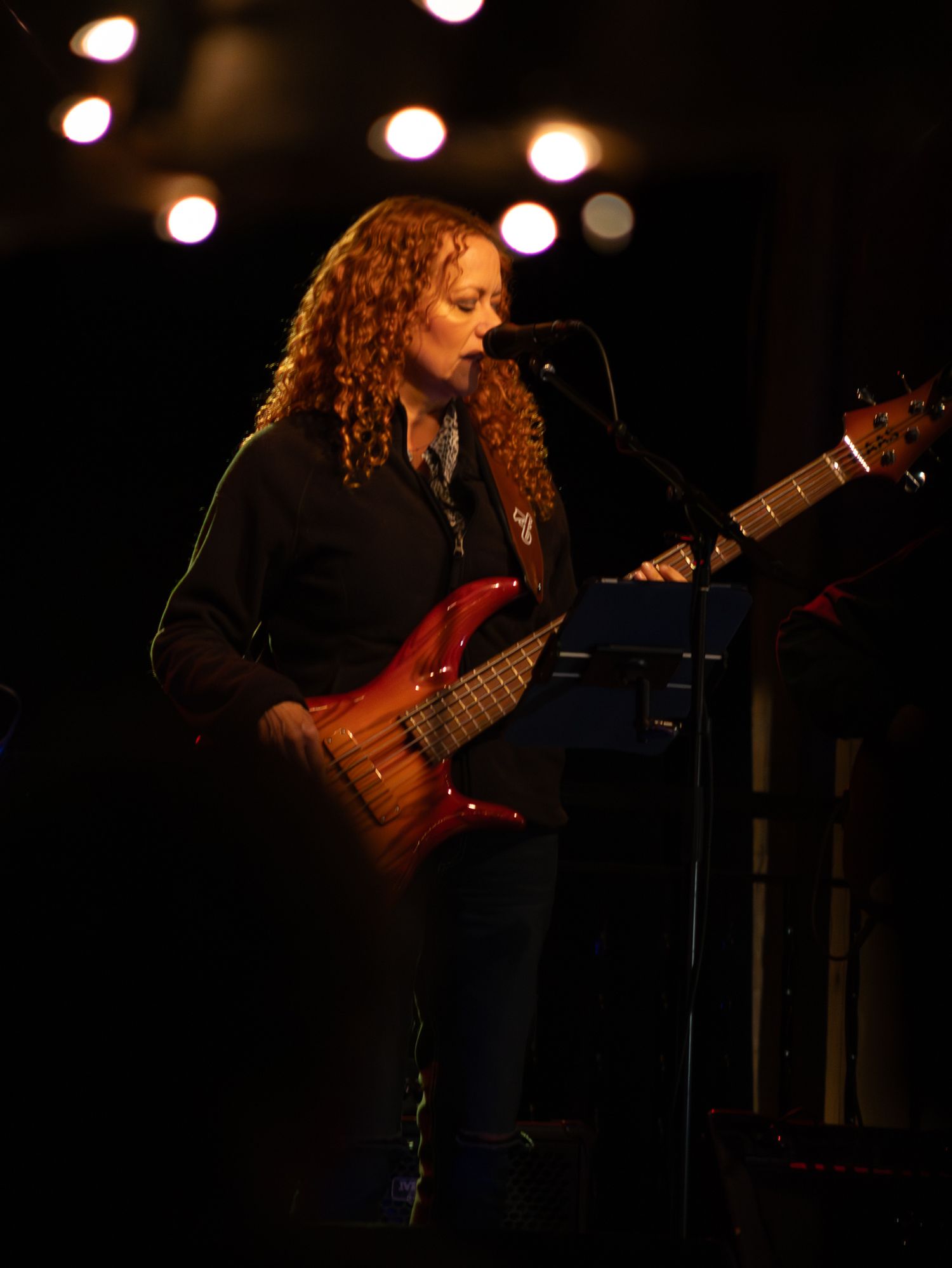 A curly-haired woman plays a red electric bass guitar on a dimly lit stage near a microphone.