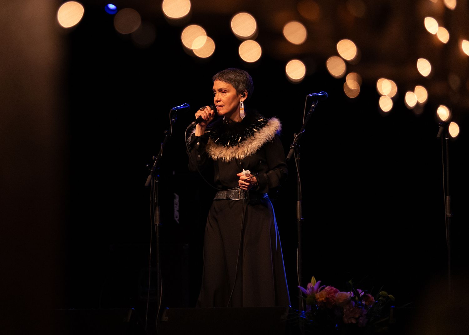A female singer performs on stage in a black outfit with fur trim, holding a microphone under warm bokeh lights.