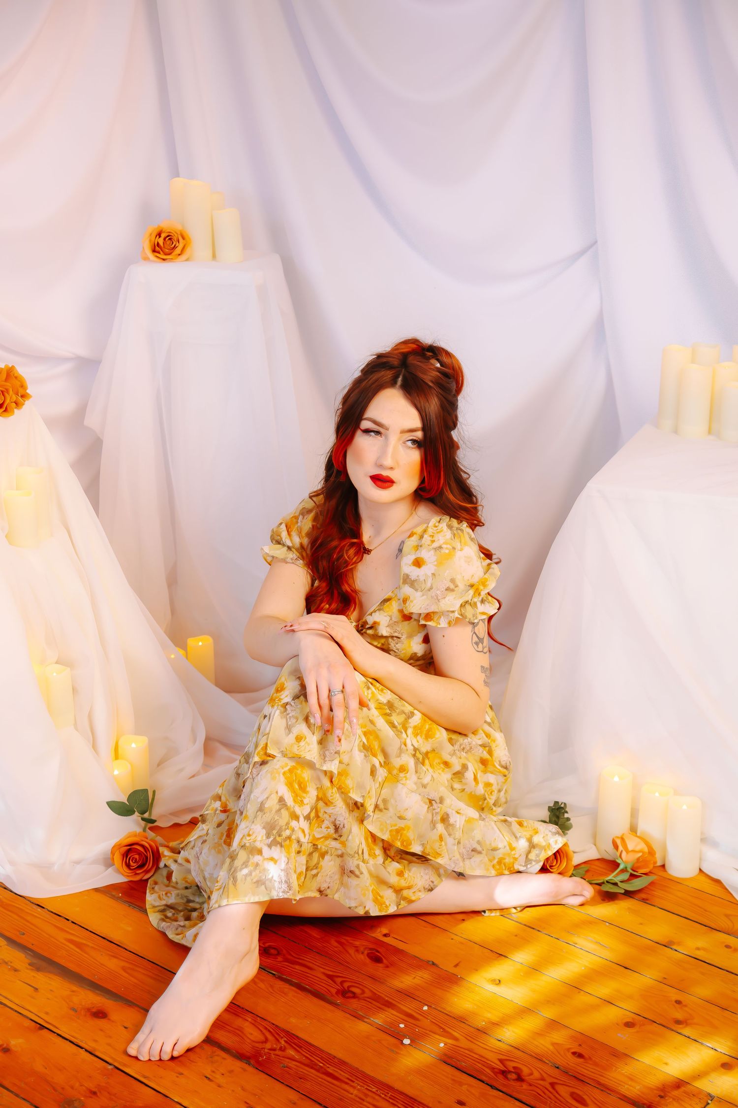 Young woman in yellow floral dress sits barefoot on wood floor surrounded by candles and orange roses against white drape backdrop.