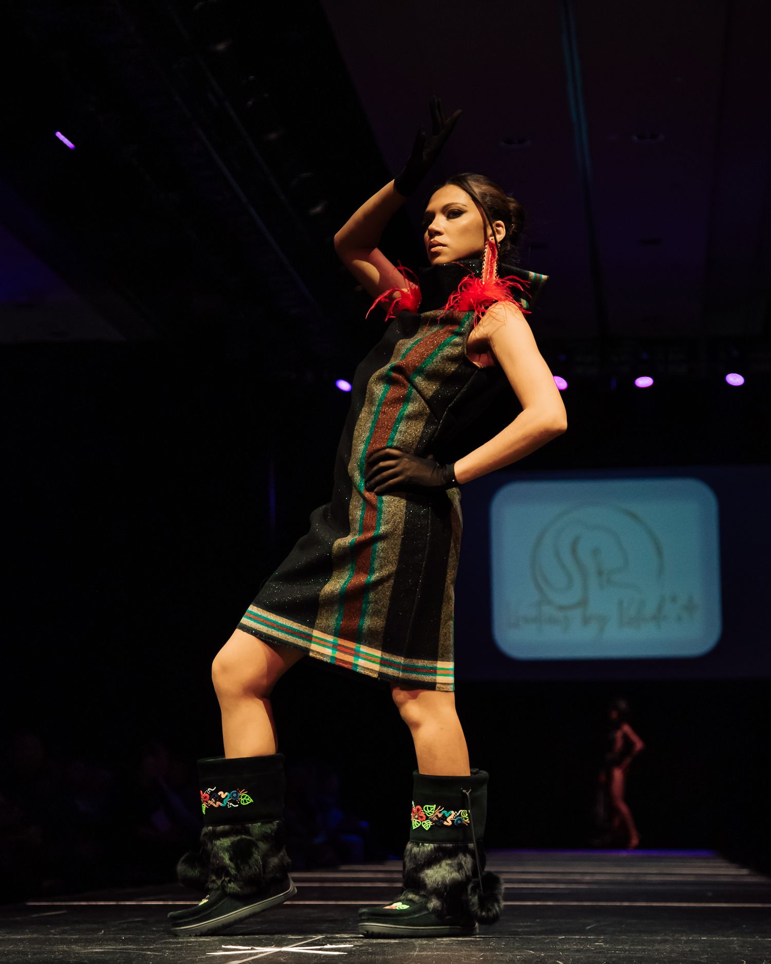 A fashion model walks the runway in a striped dress and chunky boots during a dark-lit fashion show.