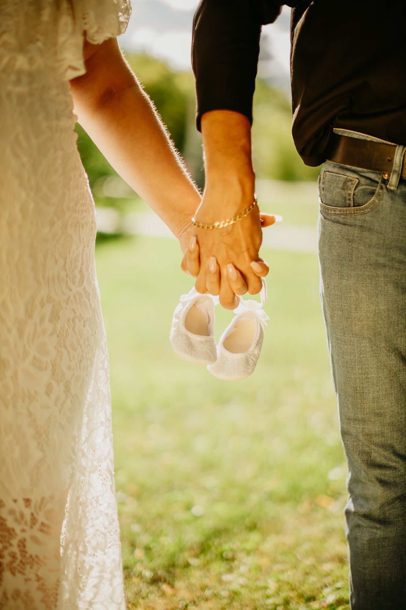 Close up of joined hands holding baby shoes against white lace dress and denim.