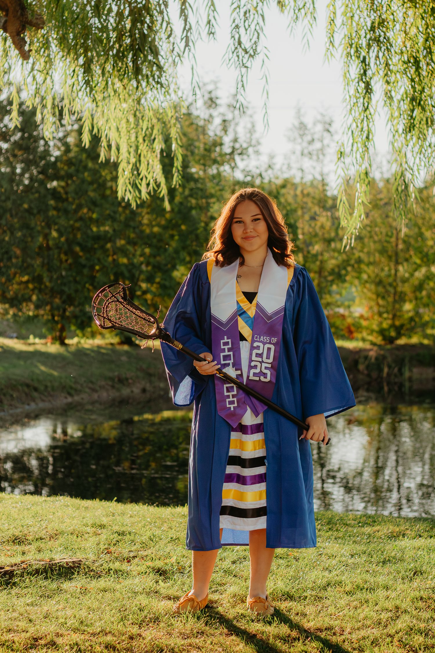 Graduate in a blue varsity jacket and purple striped dress poses by a pond under willow trees.