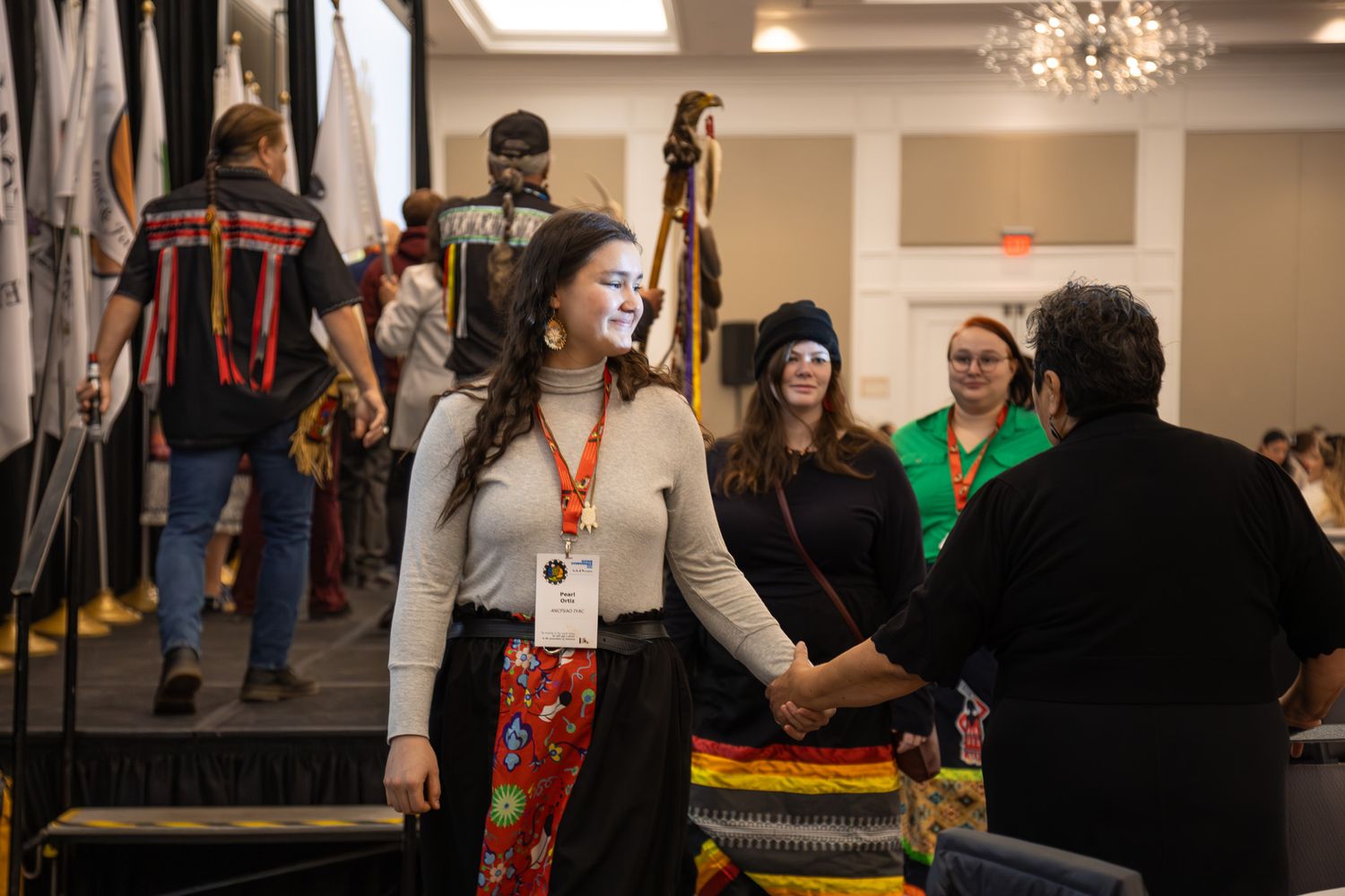 Indigenous people in traditional attire gather at a cultural conference, shaking hands and celebrating community.