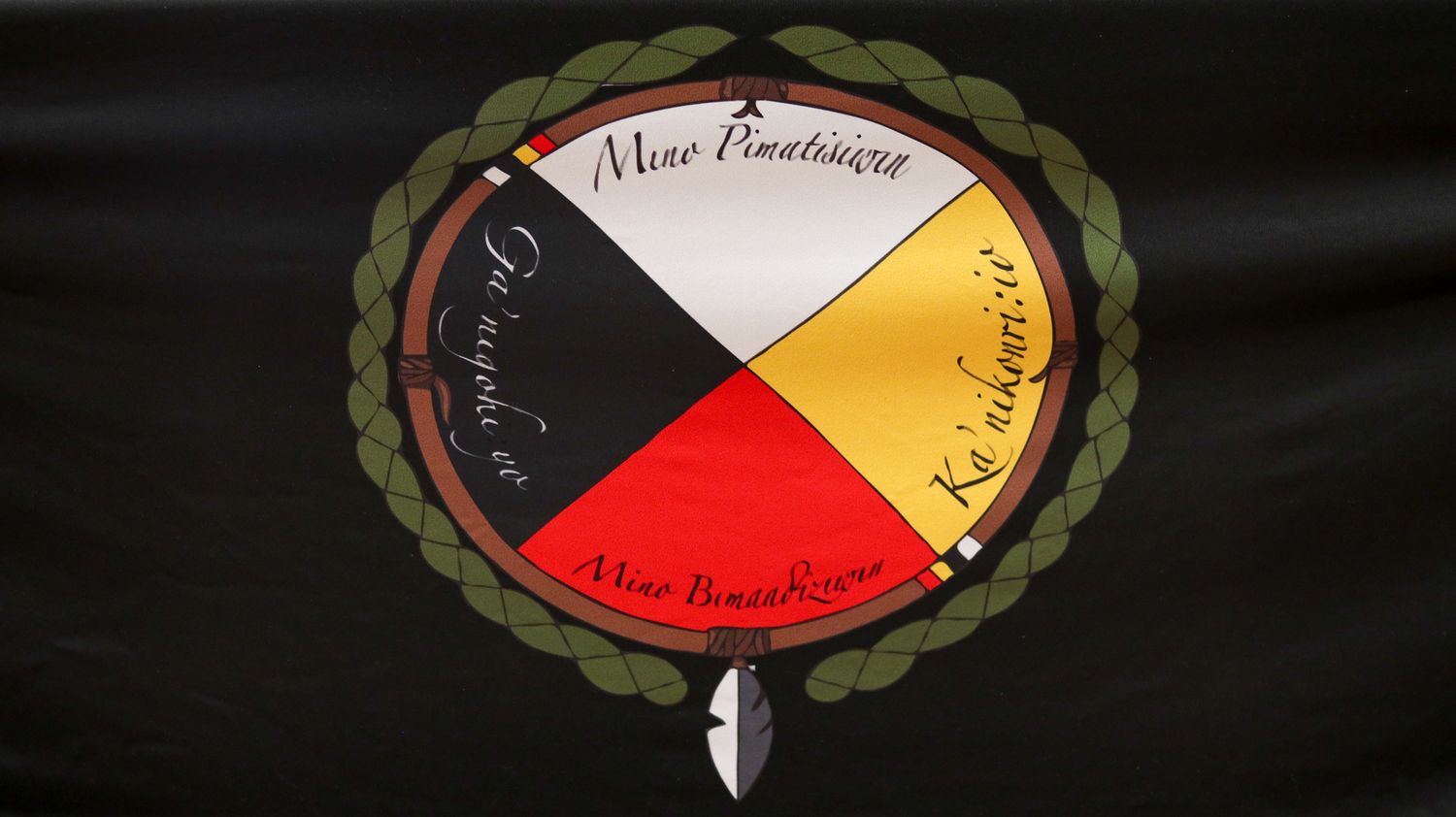 Black flag featuring a circular Indigenous emblem with red, yellow, white, and black quadrants, surrounded by a laurel wreath.