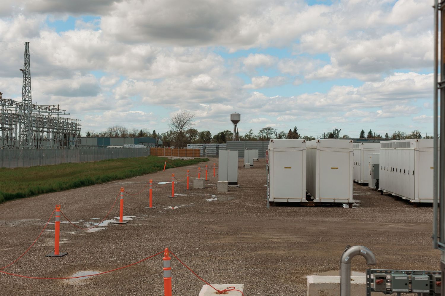 Outdoor electrical substation with white battery storage units, orange safety cones, and power infrastructure under cloudy skies.