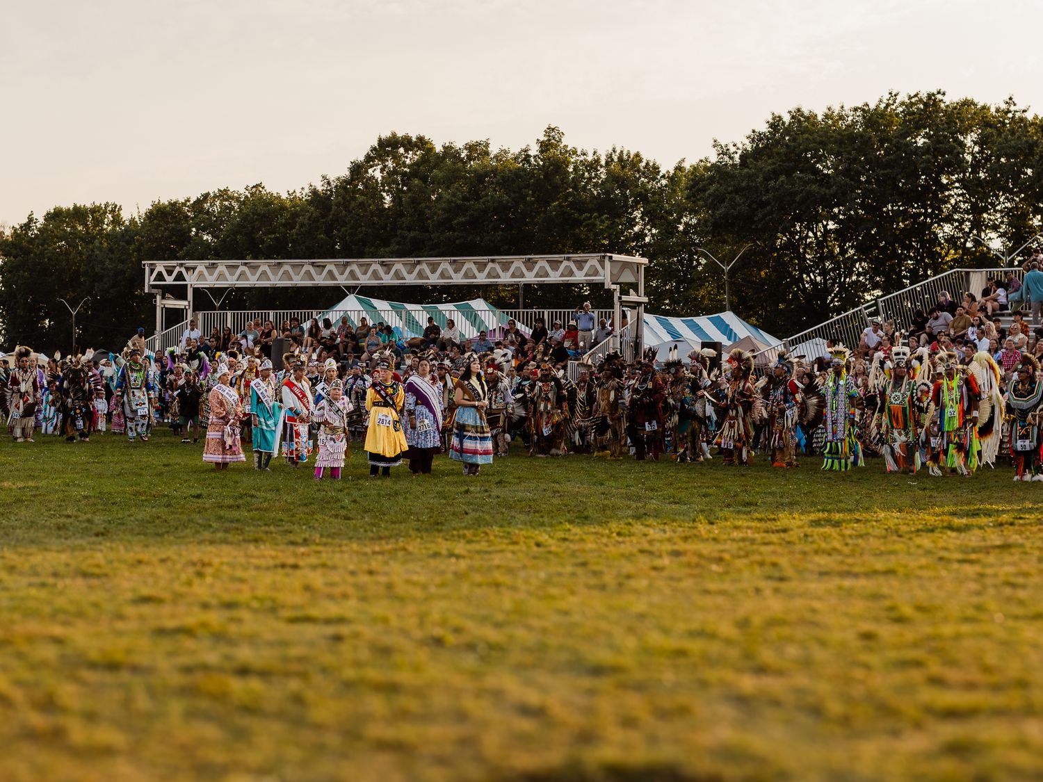 Large crowd of Indigenous dancers in colorful regalia gathered at outdoor powwow stage at dusk.