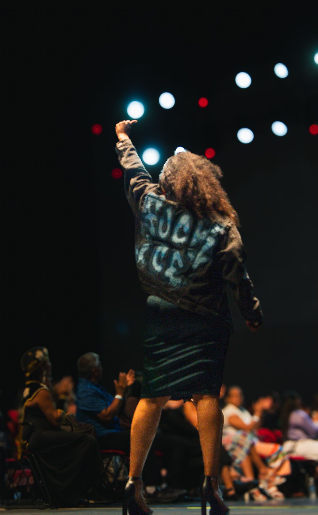 Performer on stage raising fist wearing graphic jacket and leather skirt at a live show.