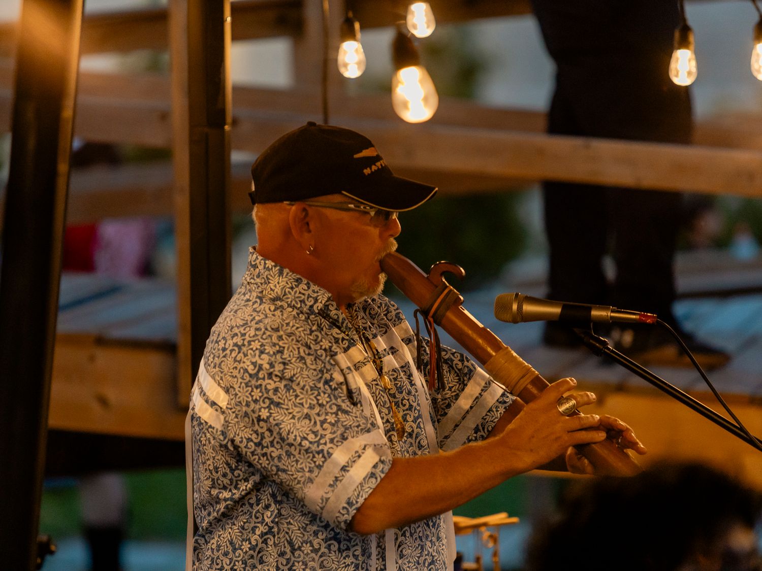 Man playing a wooden flute instrument outdoors at an evening event with warm string lights in background.