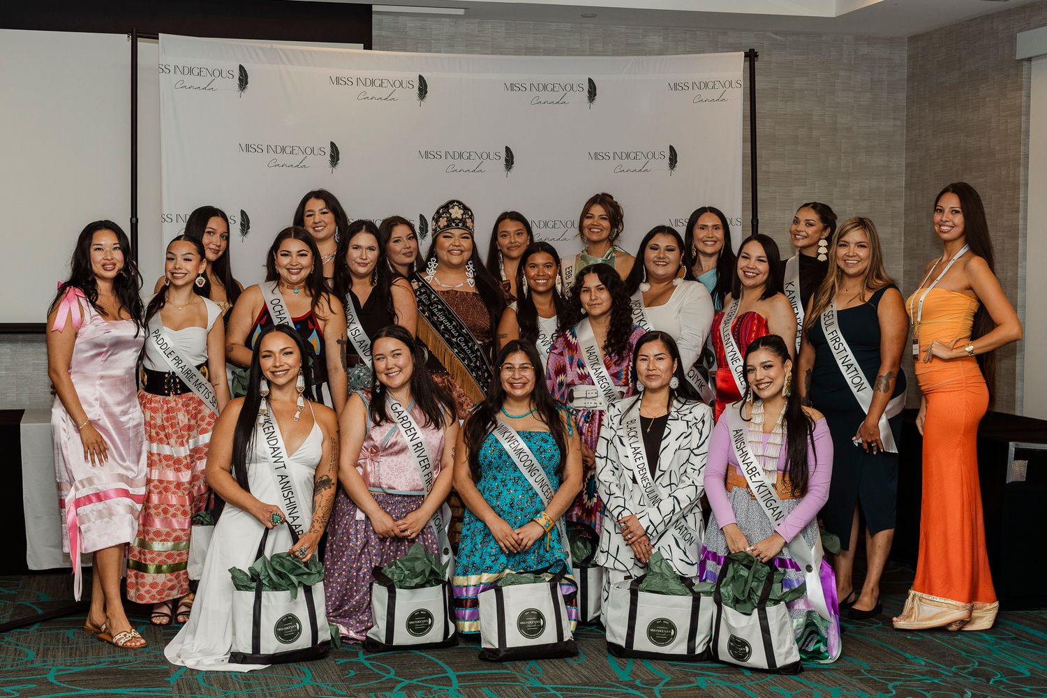 Group of Indigenous women in traditional and formal attire posing together at a pageant event backdrop.