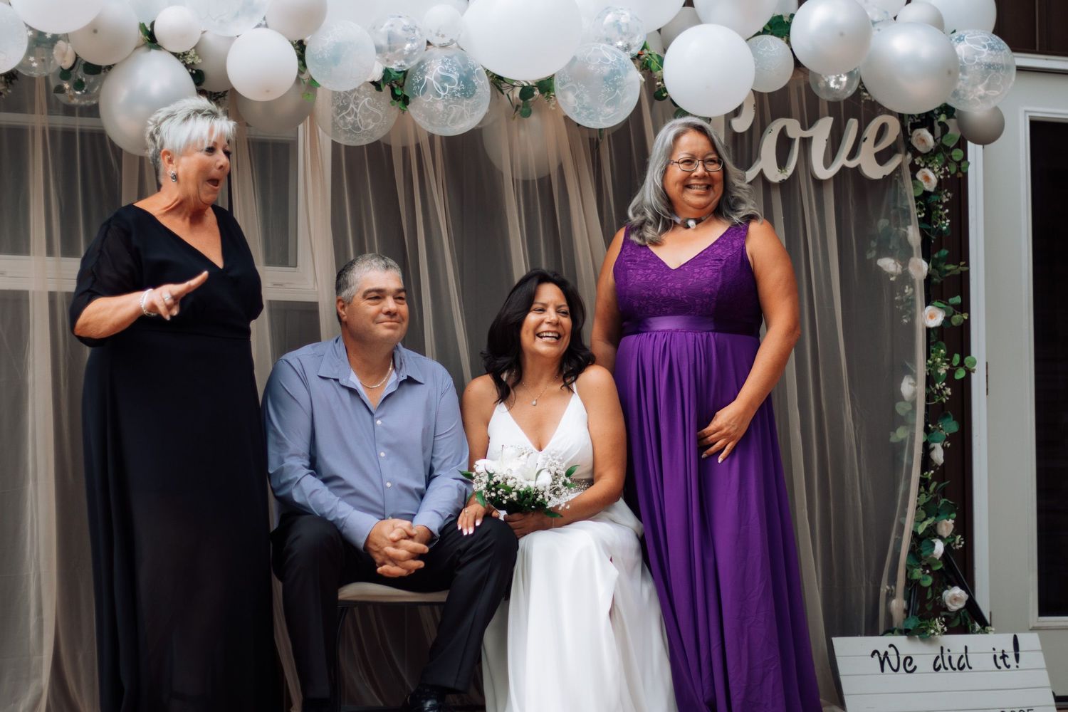 Wedding group photo with bride in white gown, bridesmaid in purple dress, under Love balloon arch.