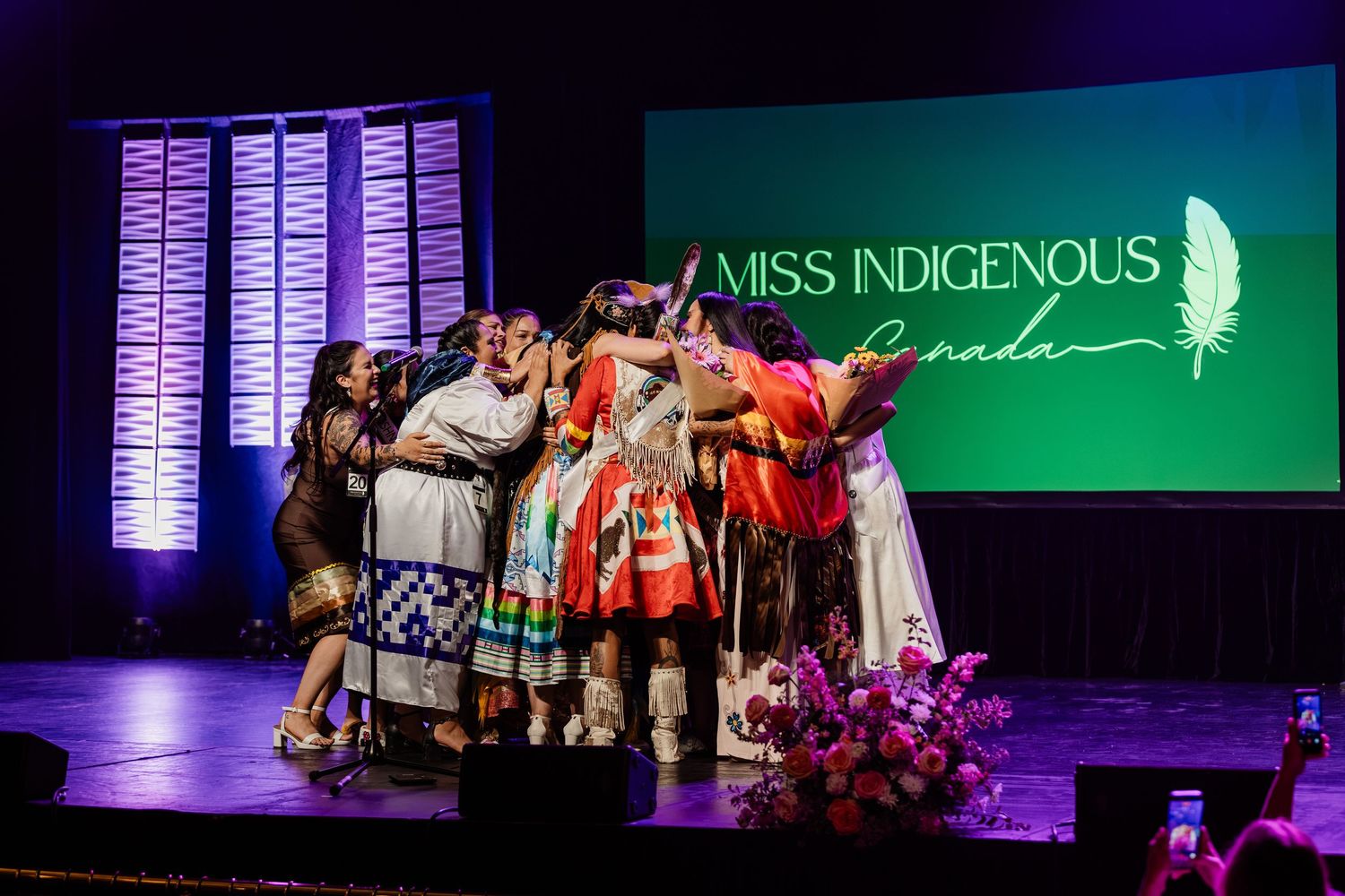 Miss Indigenous Canada pageant contestants celebrate together on stage under a colorful spotlight.