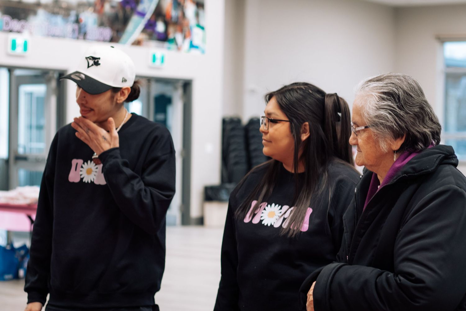 Three women in black hoodies having a conversation indoors at a community gathering space.