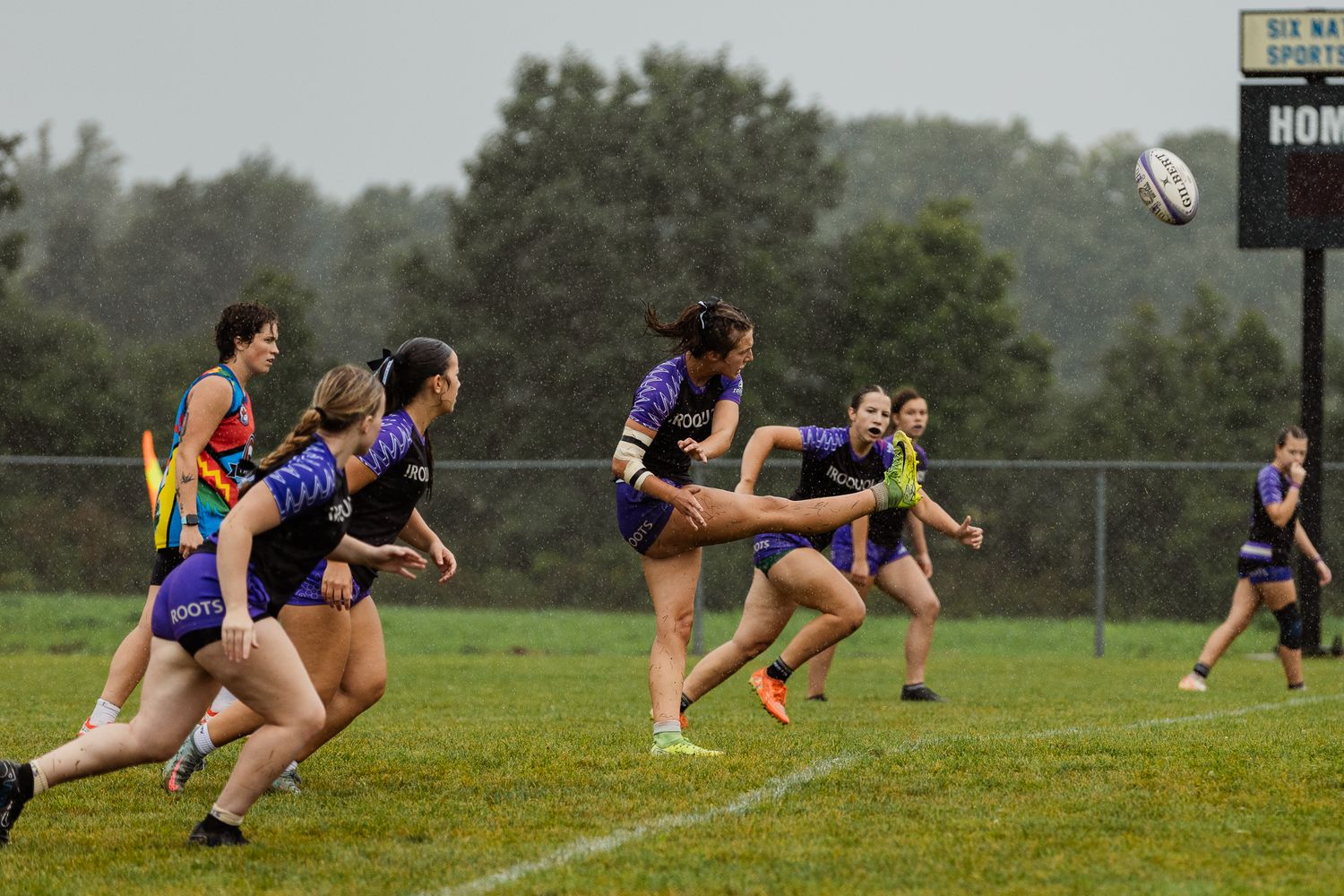 Women playing rugby on a grass field in rainy weather, with players in purple uniforms passing the ball.
