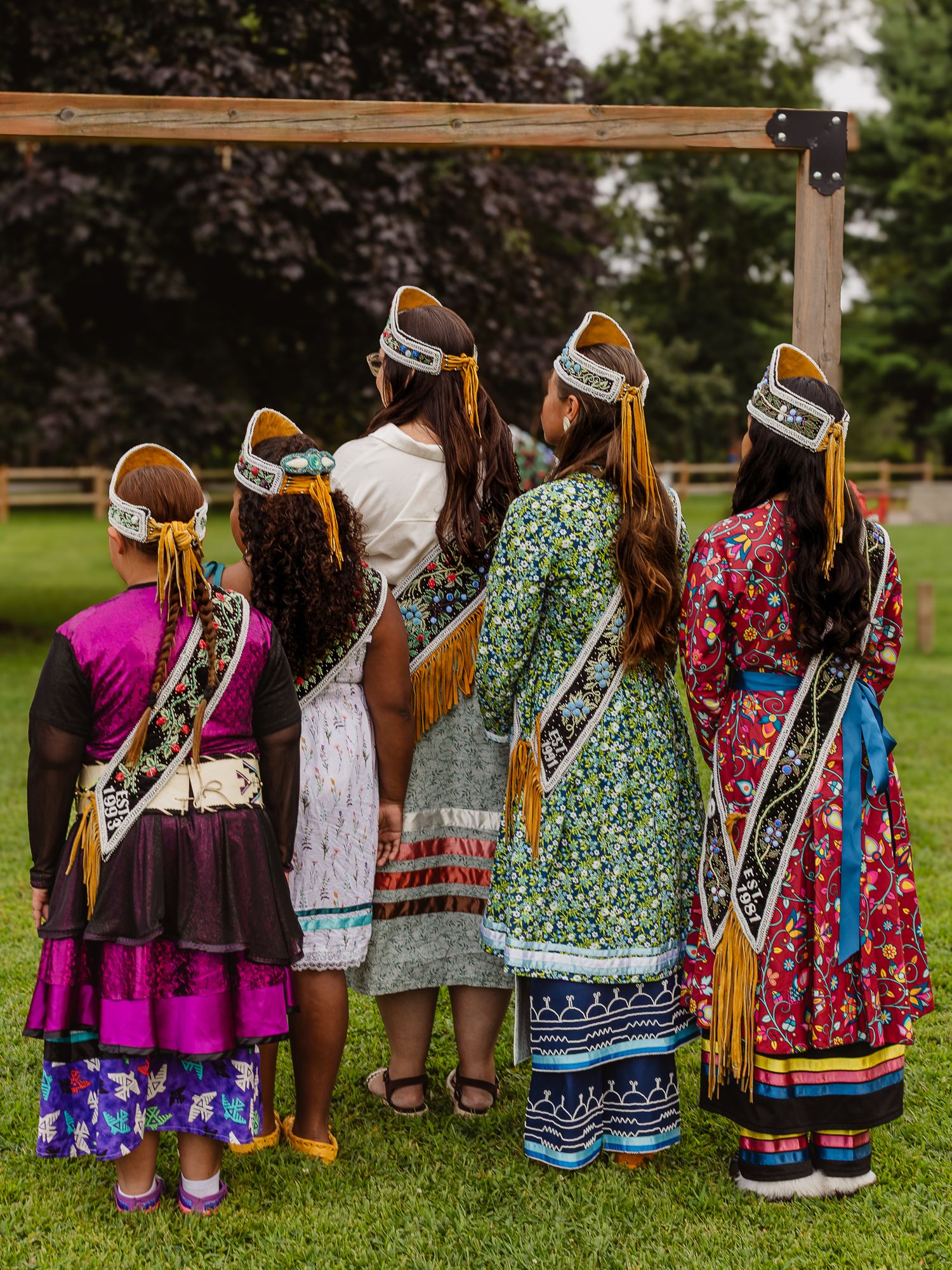 Five women in traditional Native American regalia with headbands and colorful dresses stand together outdoors.