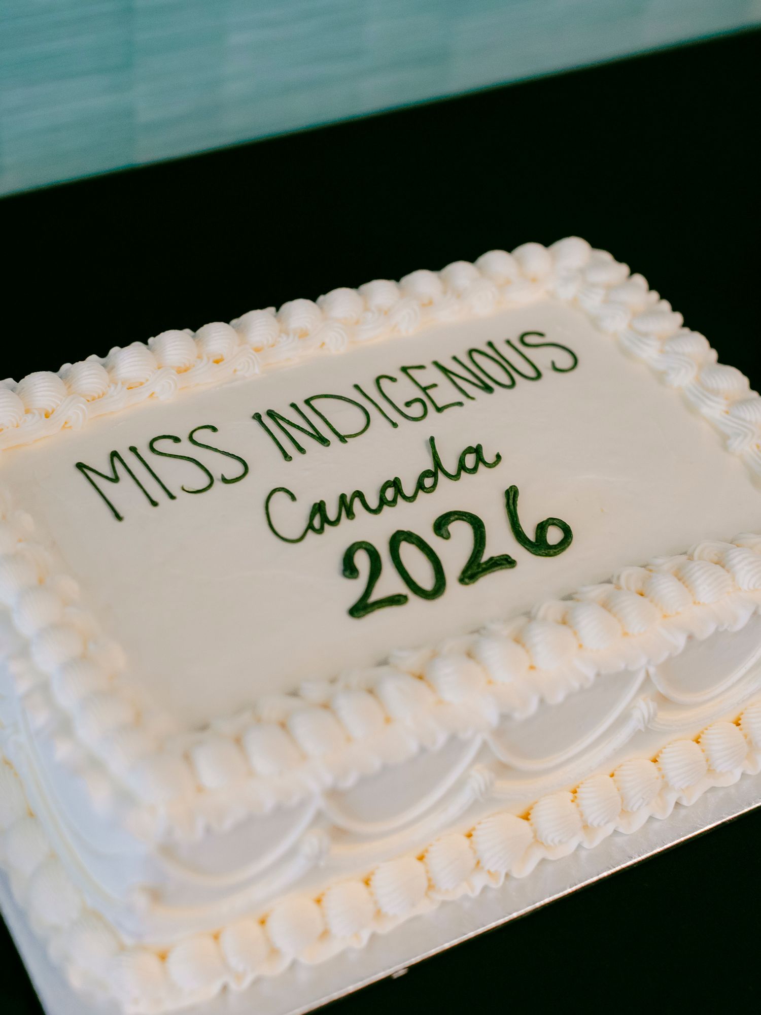 White sheet cake with green frosting text reading Miss Indigenous Canada 2026 on a dark background.