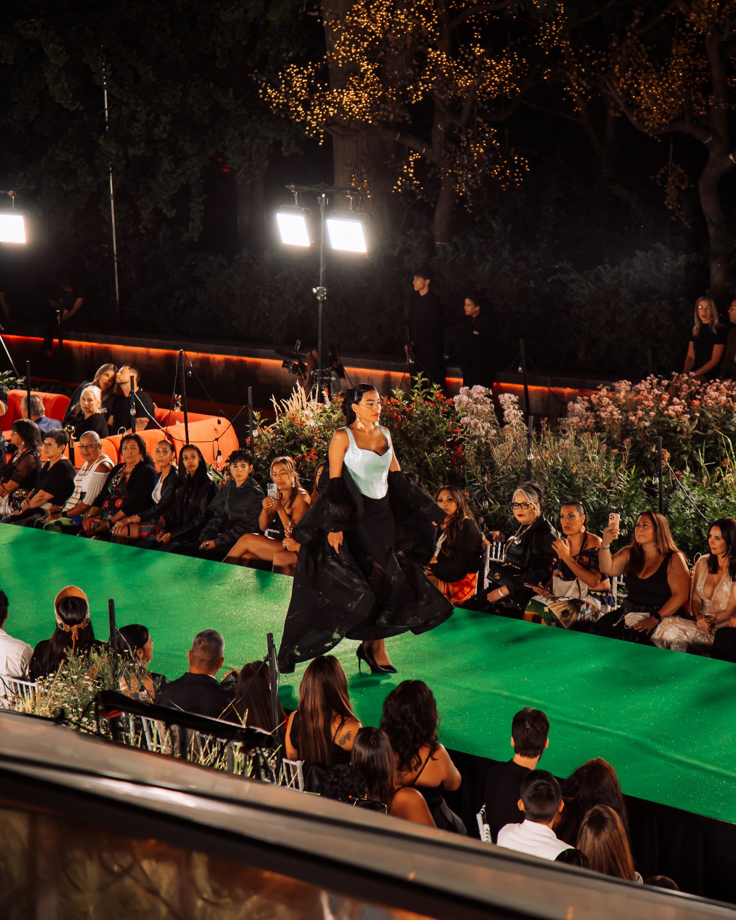 Model in white top and black skirt walks green runway at night fashion show with audience.