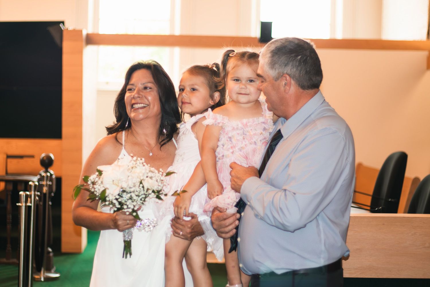 Bride in white dress holding bouquet laughs with groom and two young girls in a church wedding ceremony.