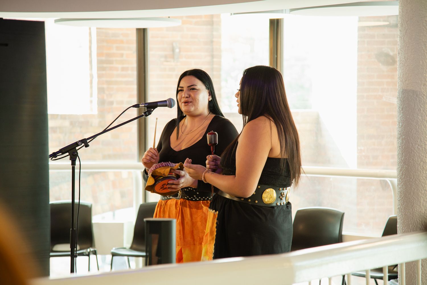 Two performers singing at microphones in front of bright windows during a daytime performance.