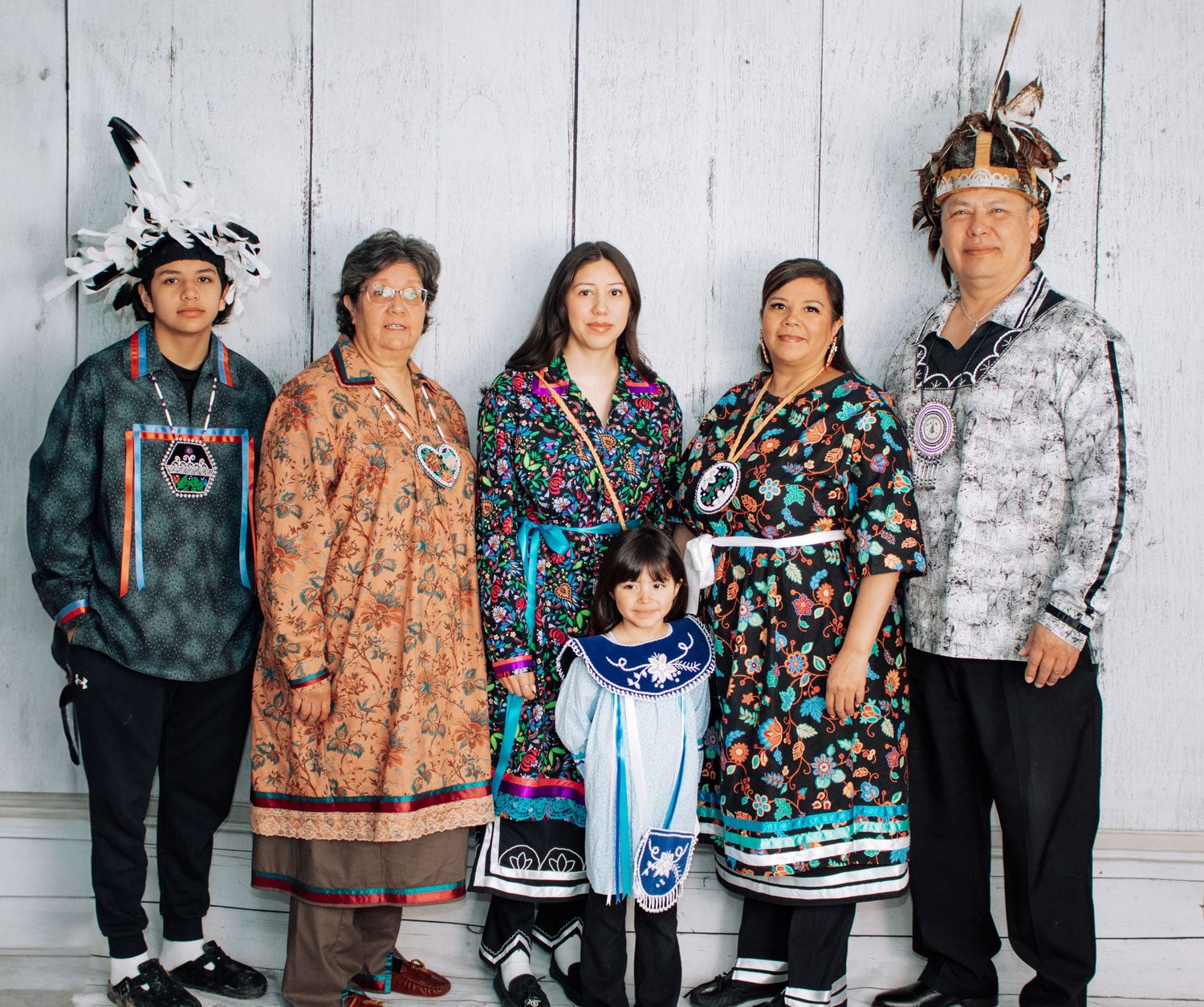 A multi-generational family wearing traditional Native American regalia poses against a white wooden backdrop.