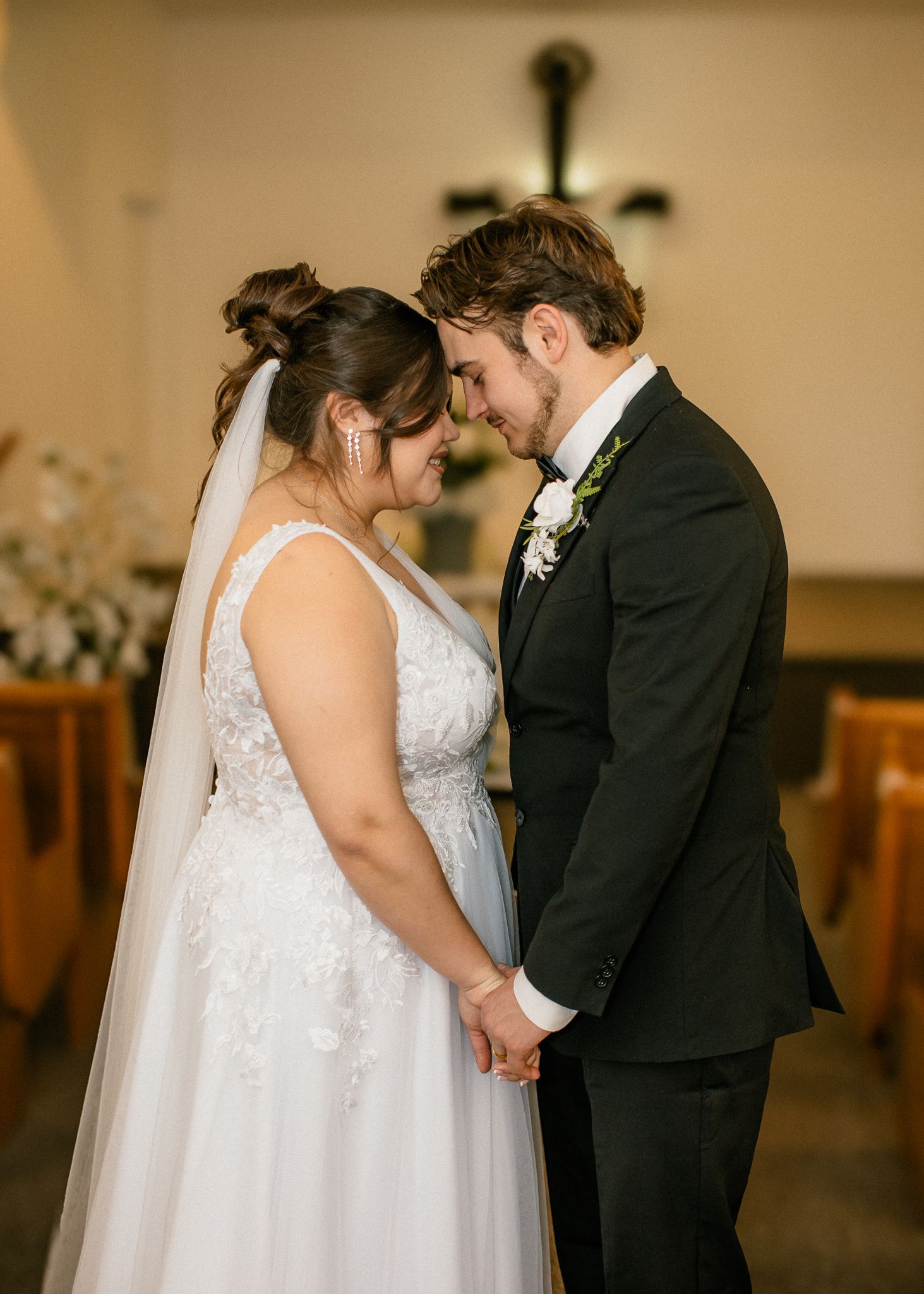 Bride in lace gown and groom in black suit hold hands and touch foreheads at church altar.