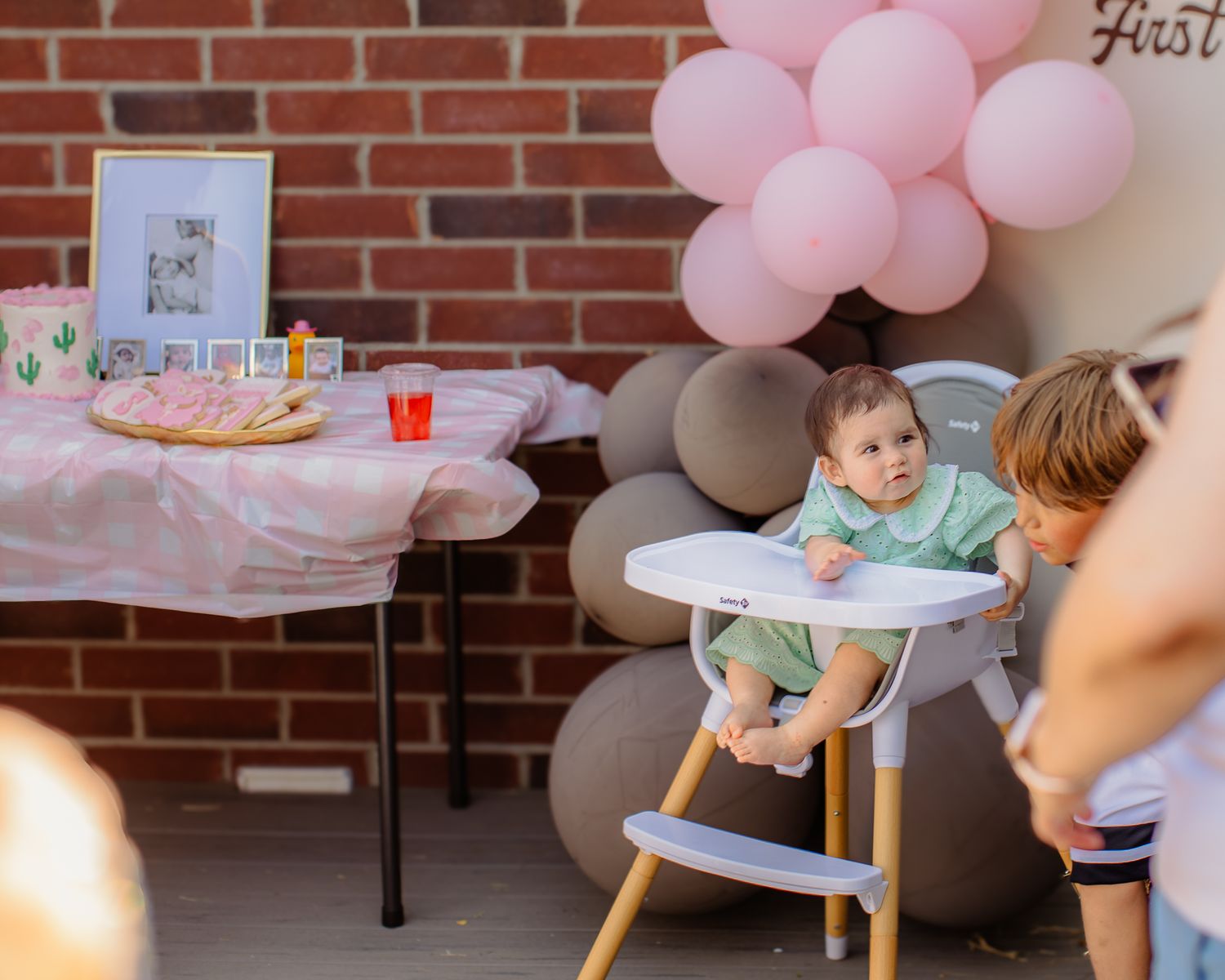 Baby in high chair at pink first birthday party with balloons and decorated table outdoors.