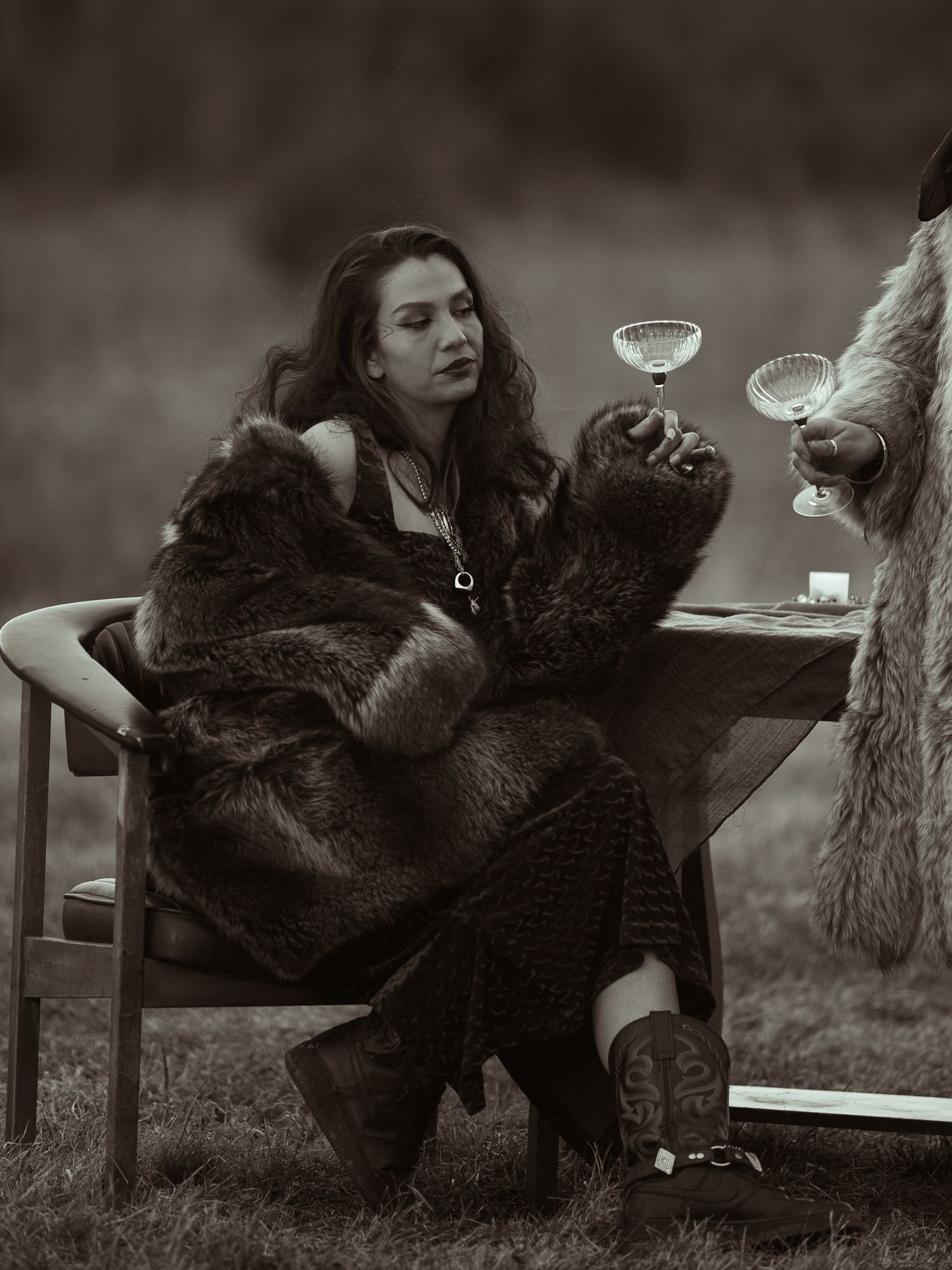 Black and white photo of woman in fur coat sitting at outdoor table with wine glasses.