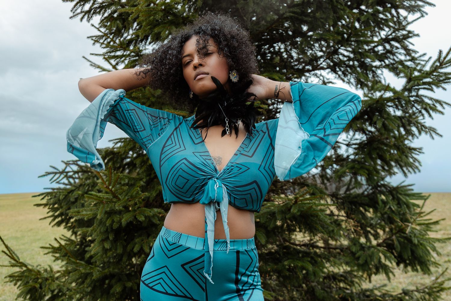 Woman in teal geometric print crop top and pants poses outdoors near evergreen tree with curly hair flowing.
