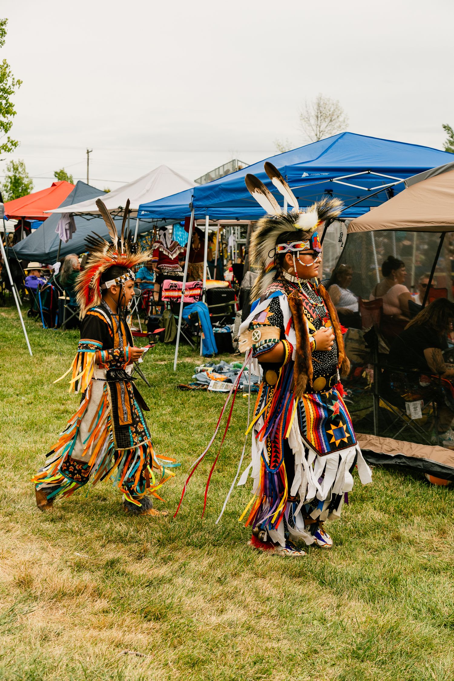 Traditional dancers in full regalia perform at an outdoor gathering with pop-up tents in background.