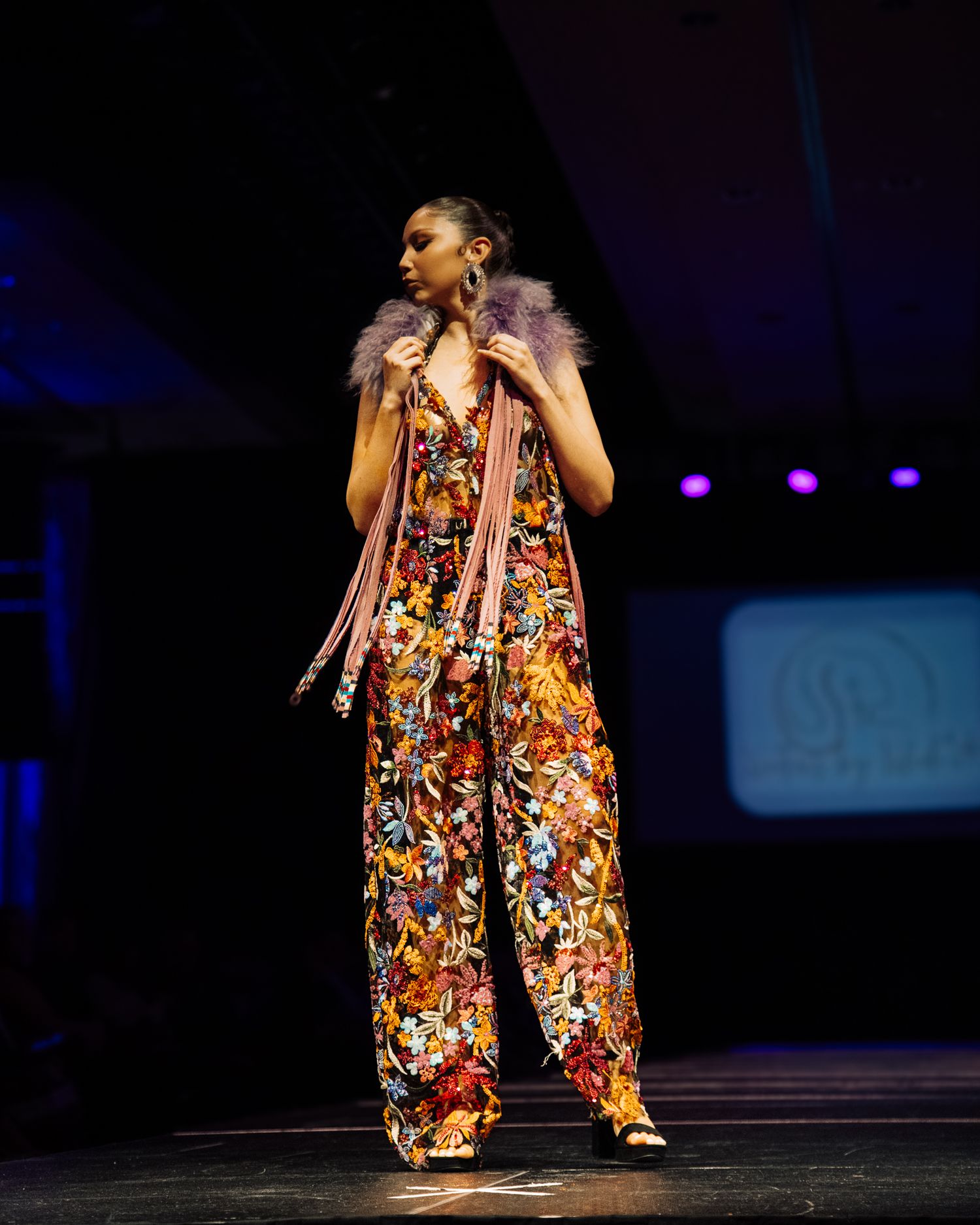 Performer in colorful floral jumpsuit dancing on dark stage with dramatic lighting.