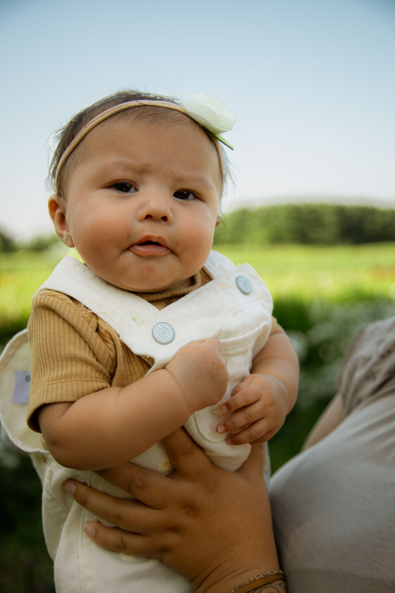 Baby wearing white and tan clothing looks curiously at camera in an outdoor setting.