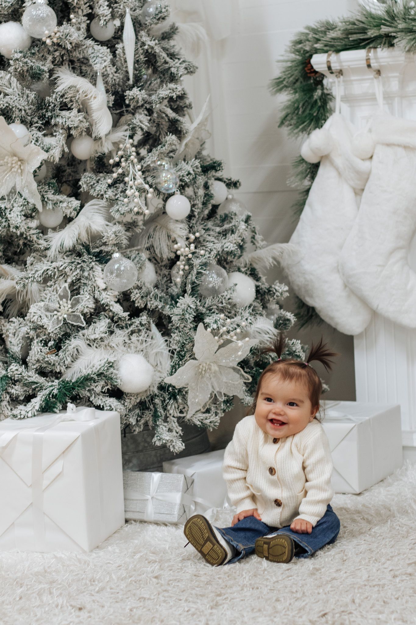 Baby sitting on white carpet by flocked Christmas tree with white decorations and wrapped presents.
