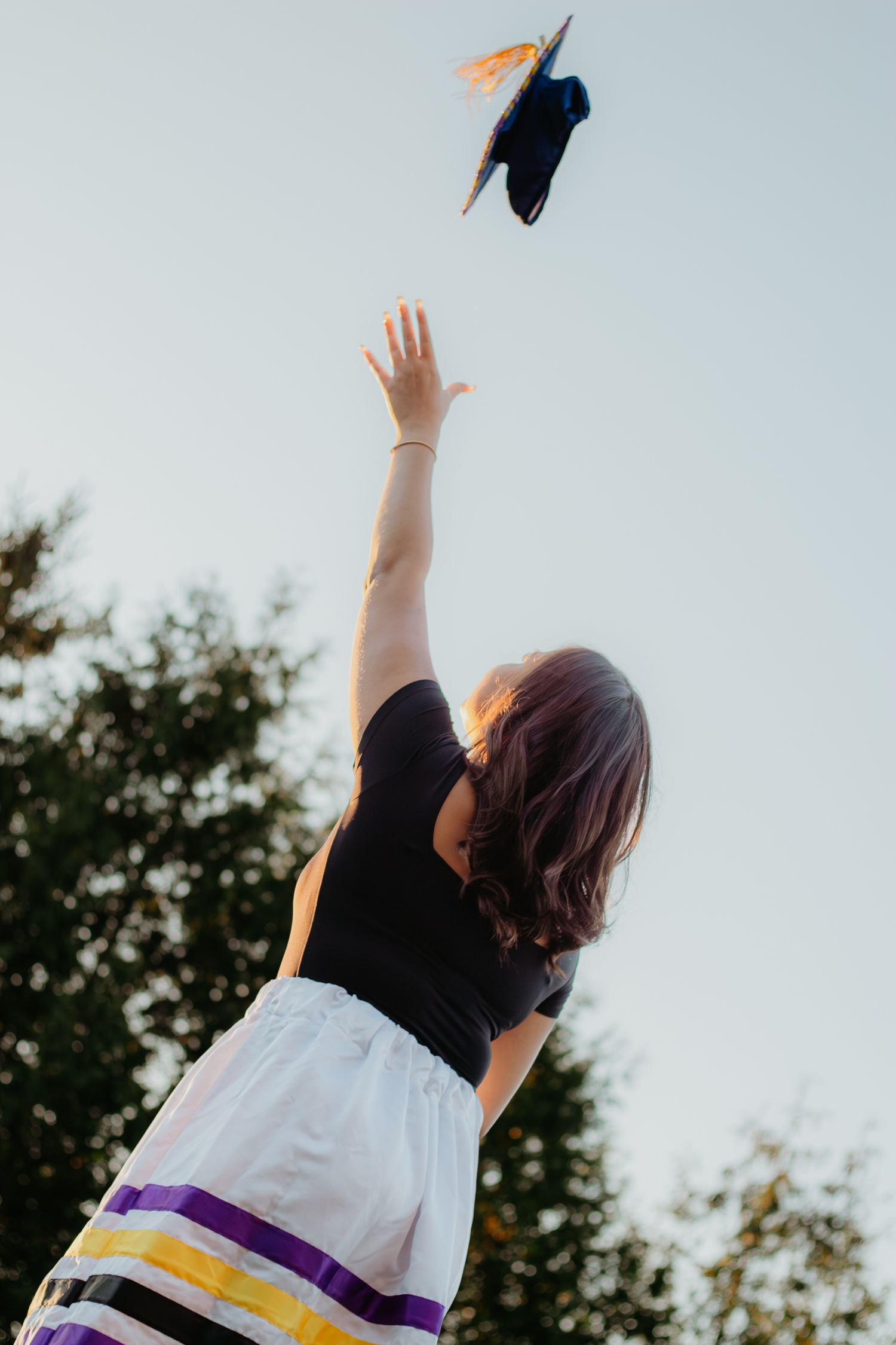 A graduate throws their cap into the air against a bright sky in celebration.