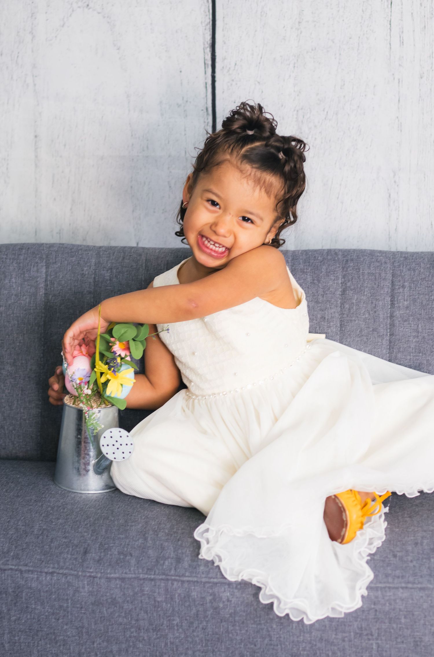 Young child in white dress posing playfully on grey couch with yellow toy.