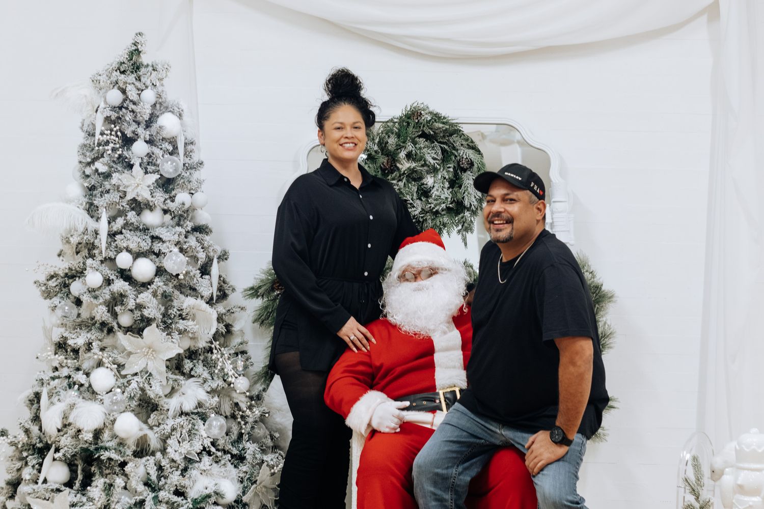 A couple poses with Santa Claus in front of a winter wonderland backdrop with flocked Christmas trees.