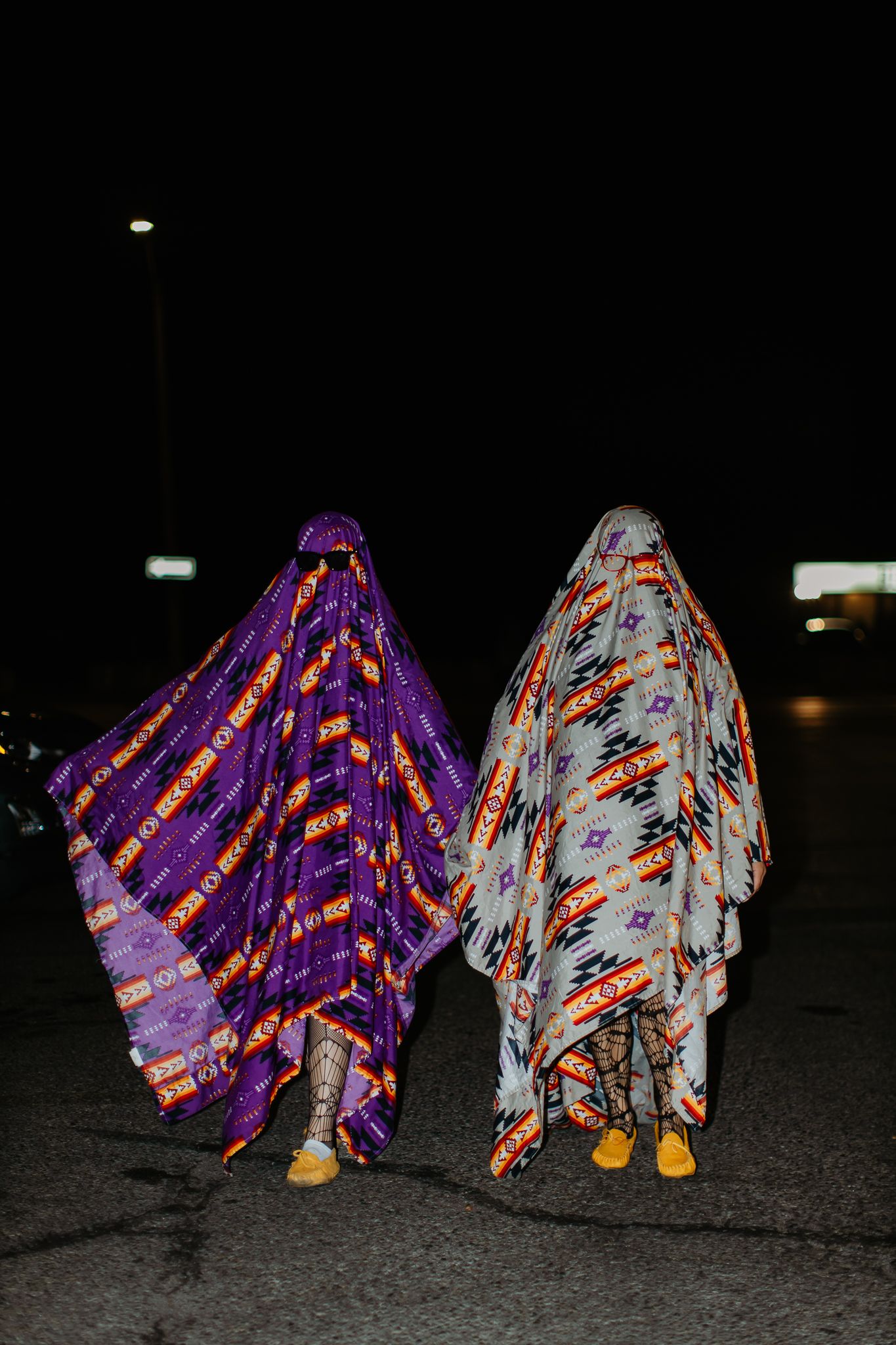 Two figures wearing colorful patterned ghost costumes standing in a dark parking lot.