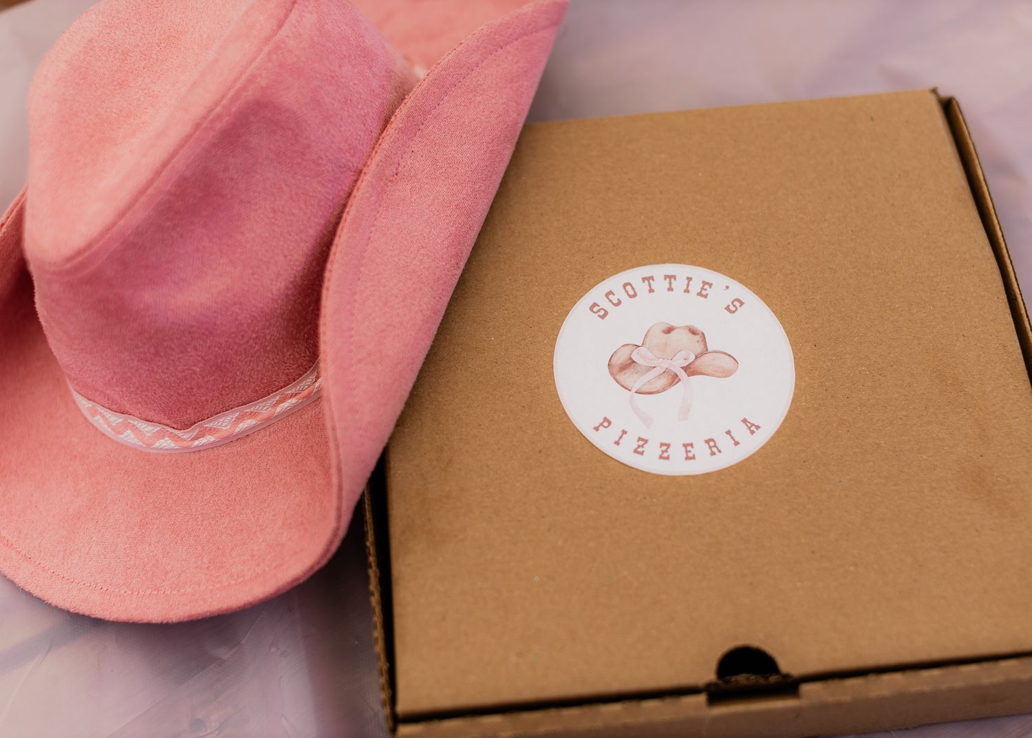 Pink cowboy hat beside a Scottie's Pizzeria branded kraft pizza box on a purple surface.